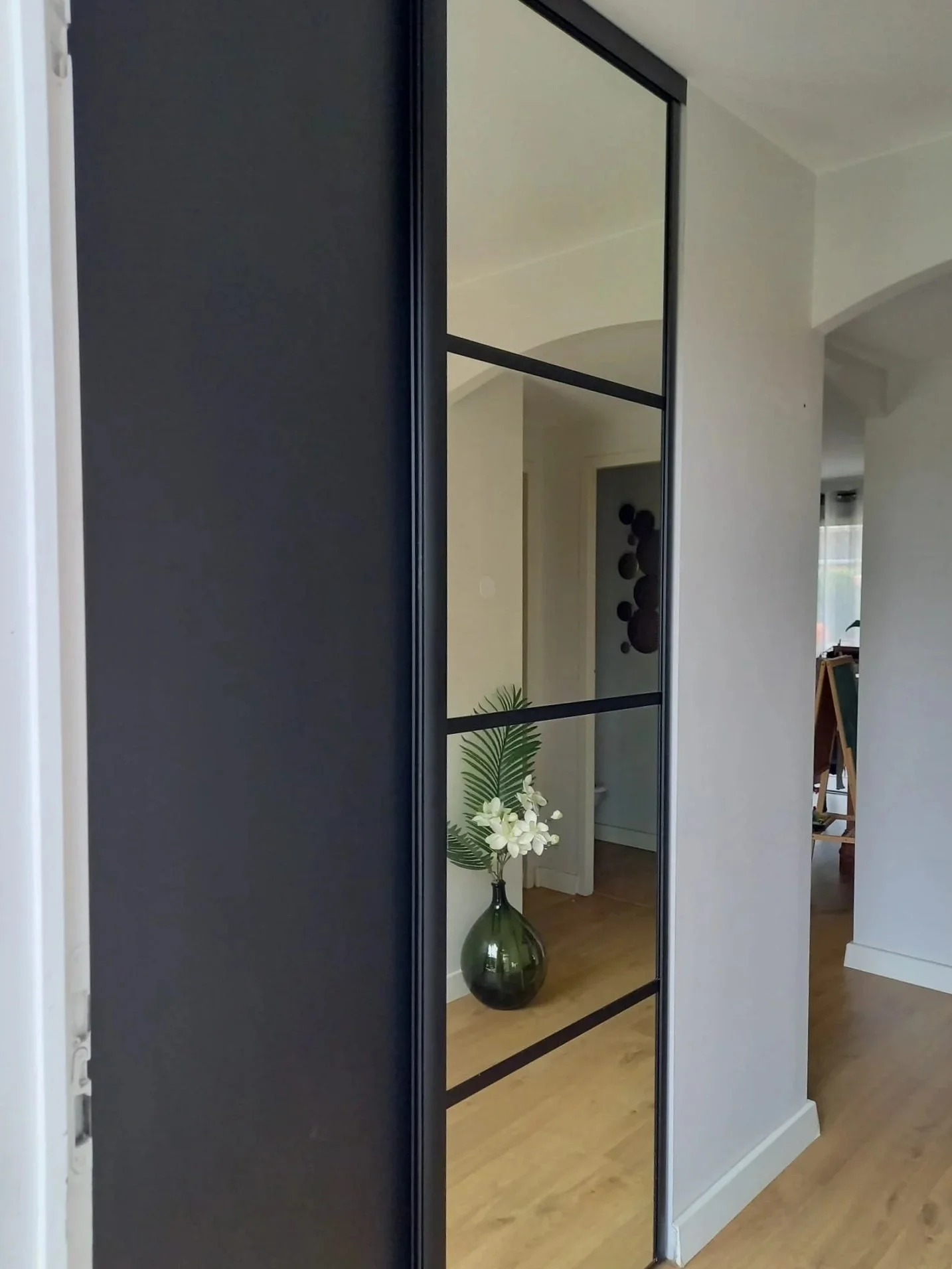 Un miroir avec une porte dans un intérieur moderne, avec un vase vert contenant des fleurs blanches et une plante verte en bas, dans une pièce avec un sol en bois.