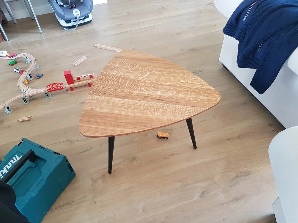 Table en bois triangulaire avec trois pattes, dans une pièce avec jouets pour enfants, notamment un circuit de train, un camion jouet orange, et un coffre à outils bleu. On voit aussi un canapé avec un vêtement bleu posé dessus.
