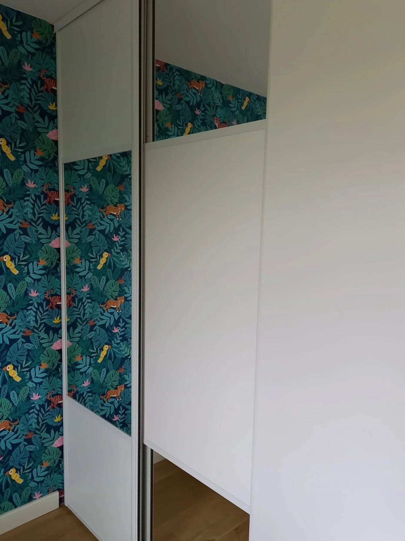Armoire blanche avec des portes coulissantes, une à motifs tropicaux avec des feuilles, des tigres, des toucans et des oiseaux colorés, autre porte blanche sans motif visible, sol en bois, mur blanc.