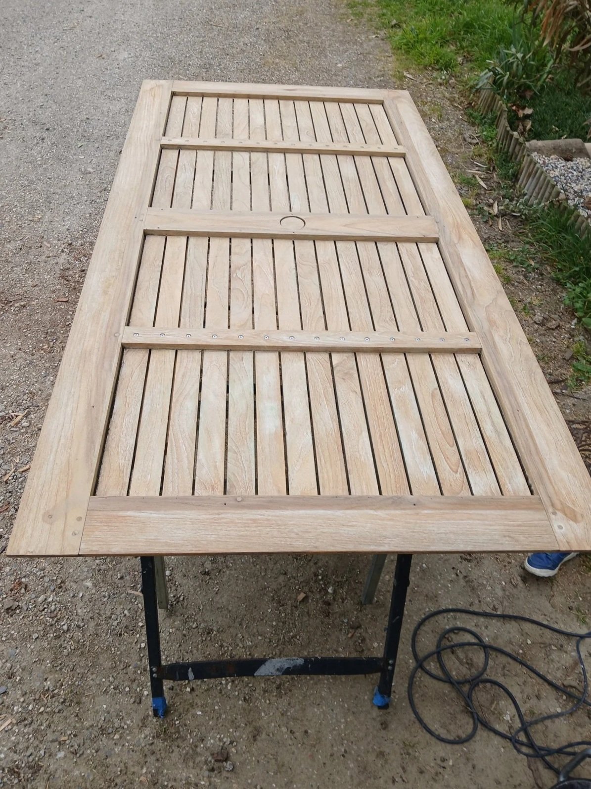 Table en bois en cours de fabrication, posée sur un support métallique, avec un cordon électrique à côté, située à l'extérieur sur un sol en gravier avec des plantes en arrière-plan.