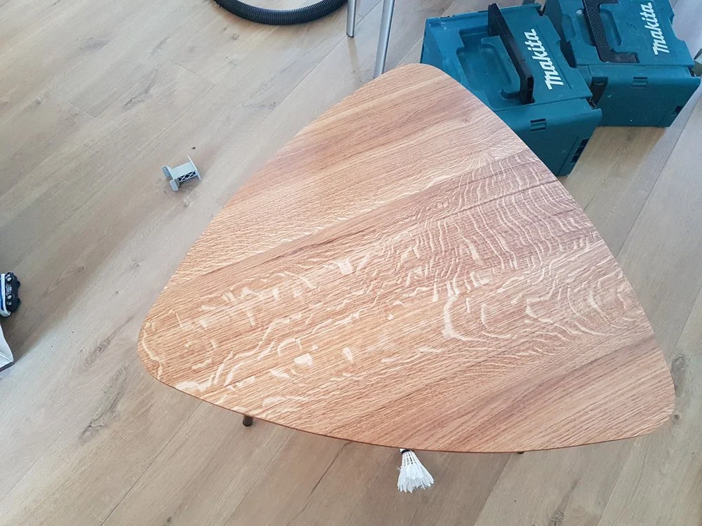 Table en bois triangulaire sur un sol en bois clair, avec deux boîtes à outils Makita en arrière-plan.
