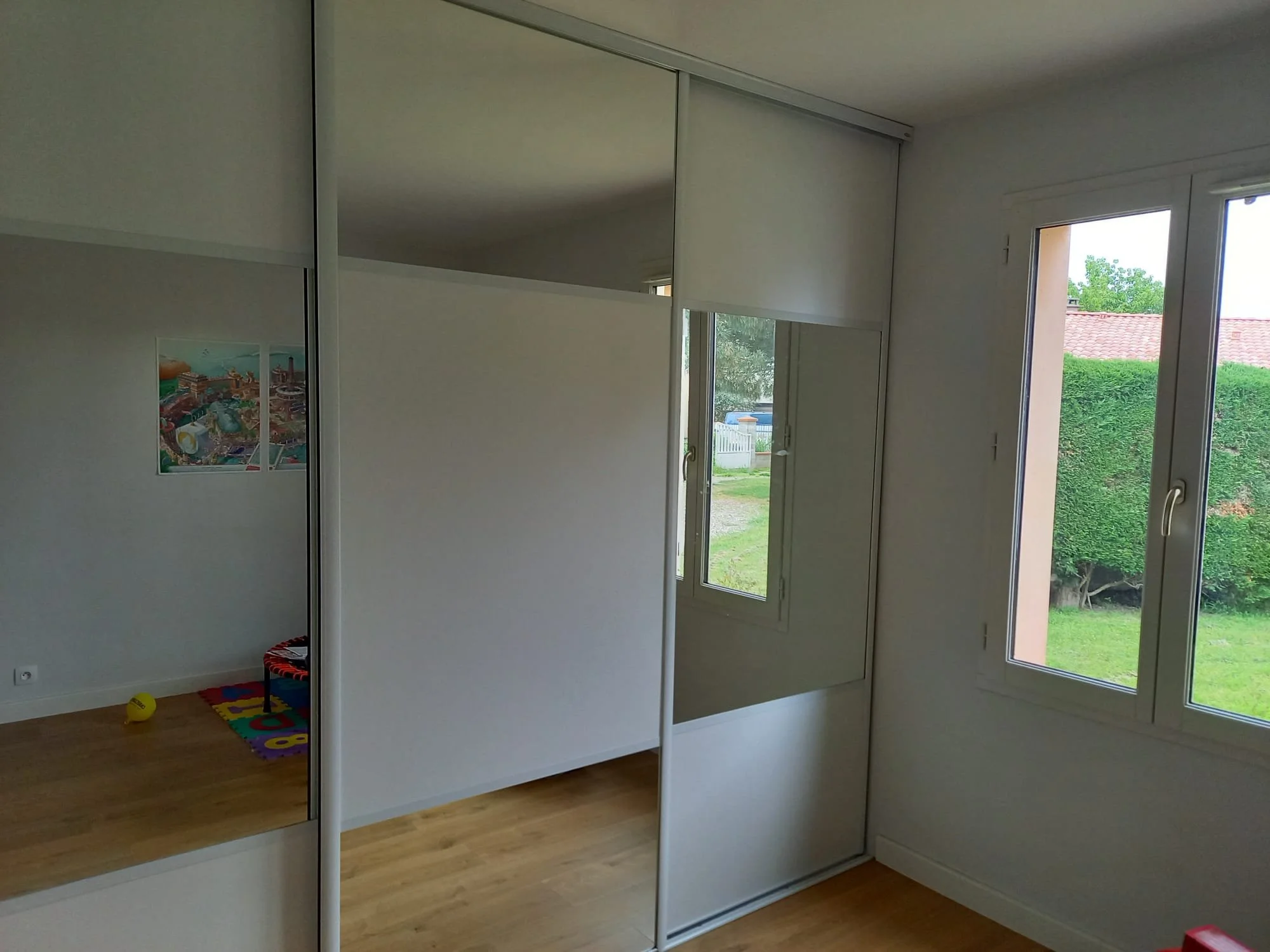 Une chambre avec de grandes fenêtres donnant sur un jardin verdoyant, un placard avec portes miroir, un sol en bois clair.