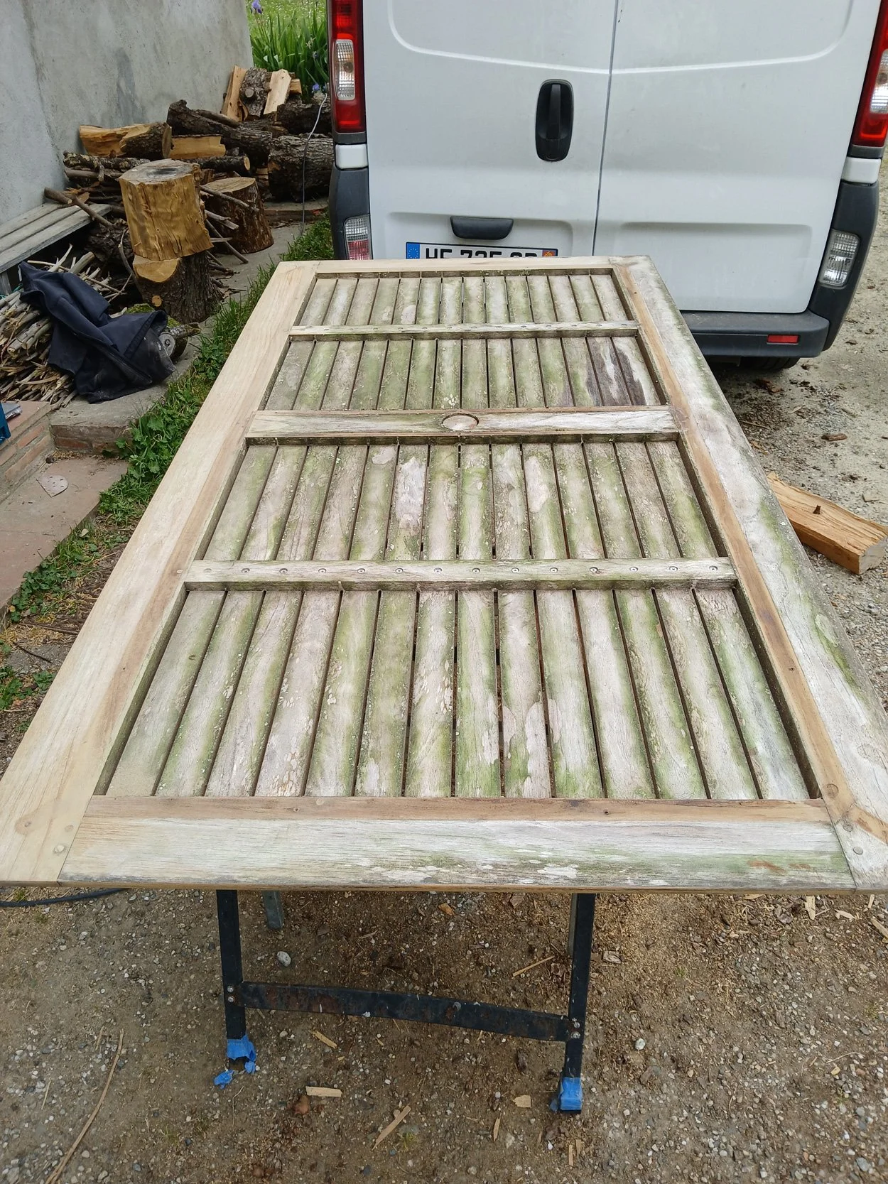 Table en bois en cours de rénovation avec une surface en planches en bois usées et une structure en bois clair, située à l'extérieur sur un support métallique, près d'une voiture blanche et de bois de chauffage.