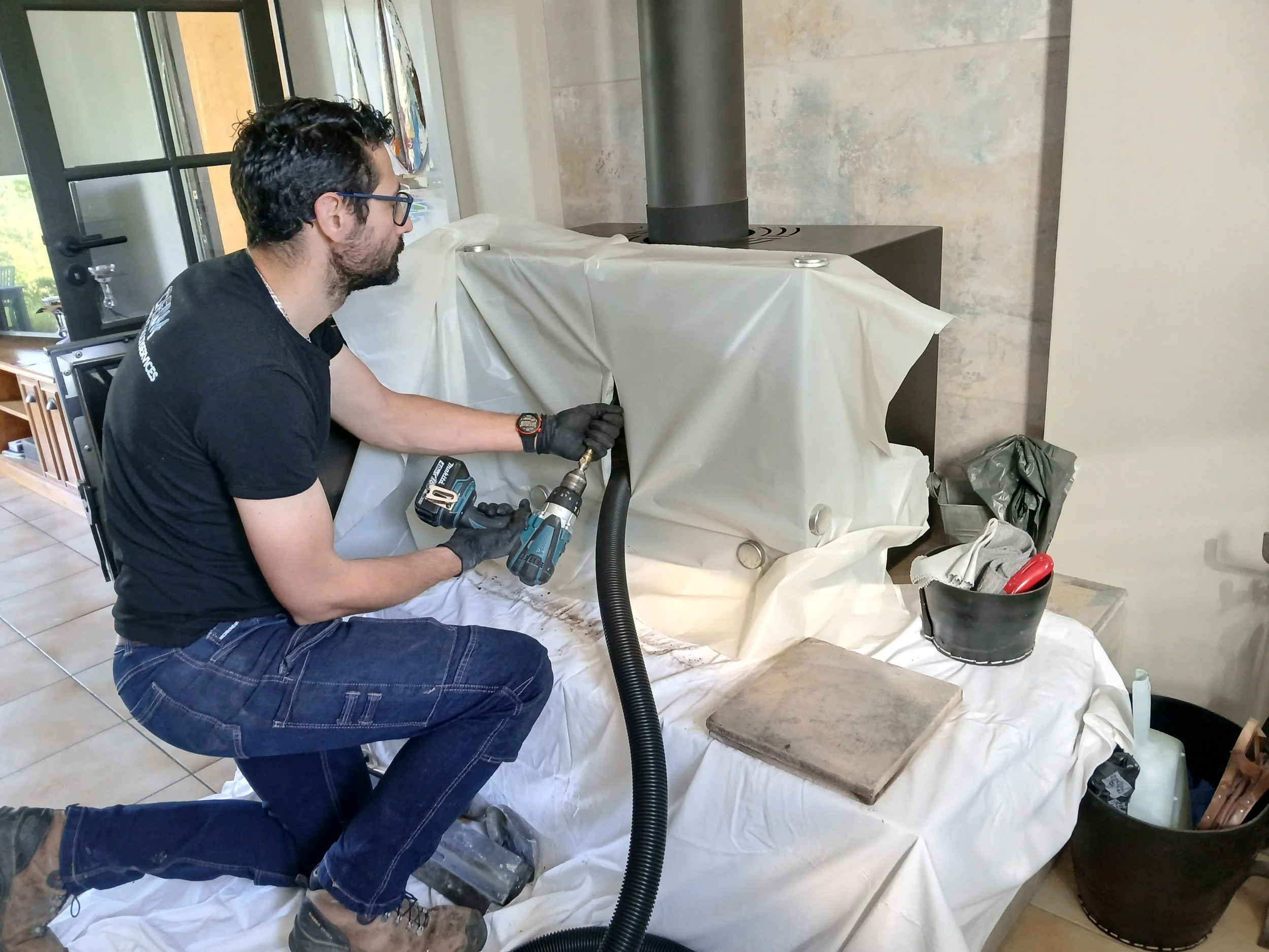 Jeune homme en jeans et t-shirt noir, portant des gants, utilisant une perceuse sur un meuble recouvert d'une bâche en plastique dans une pièce intérieure.