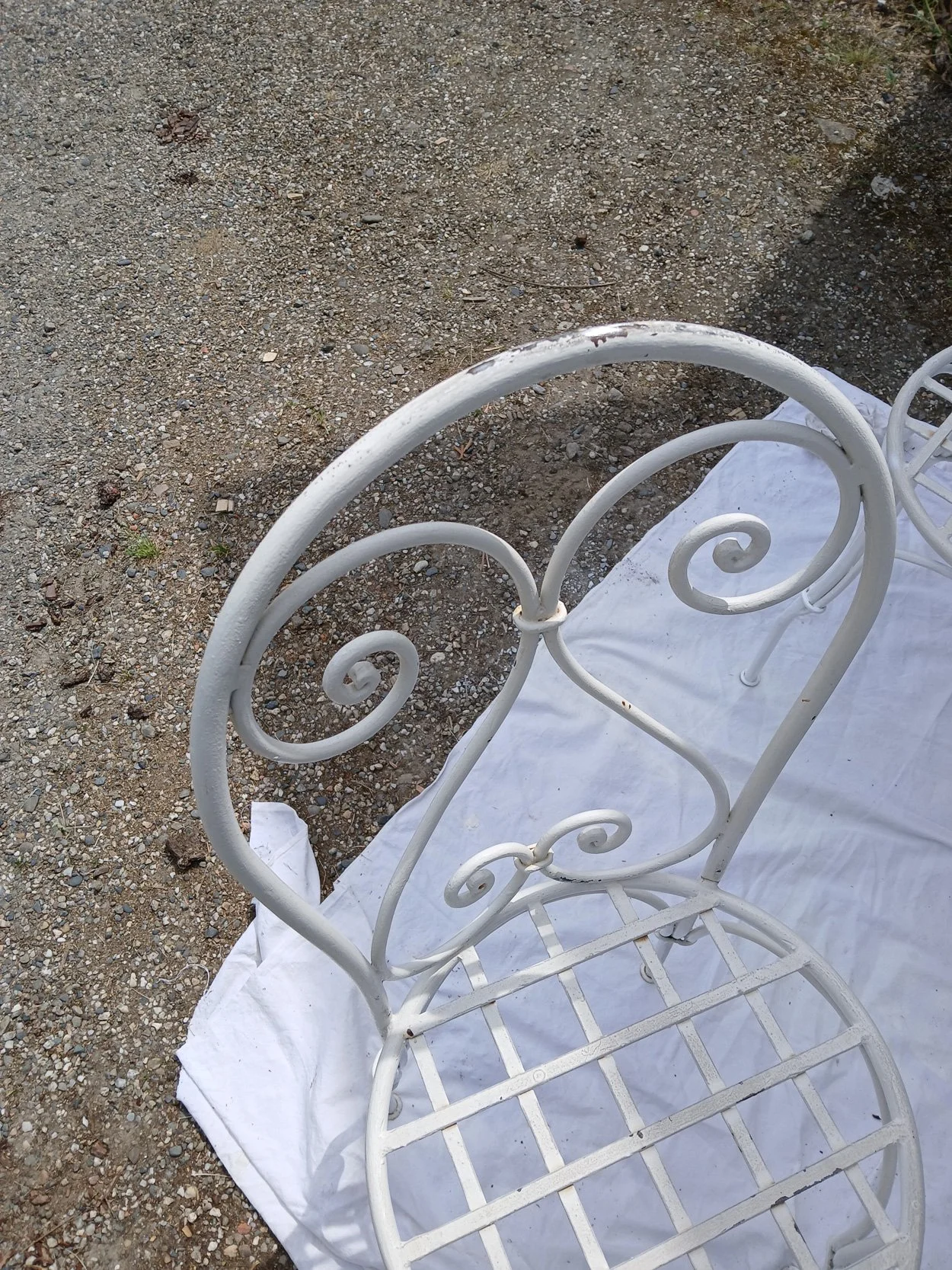 Une chaise en métal blanche avec des motifs en spirale, placée sur un drap blanc sur un sol en gravier.