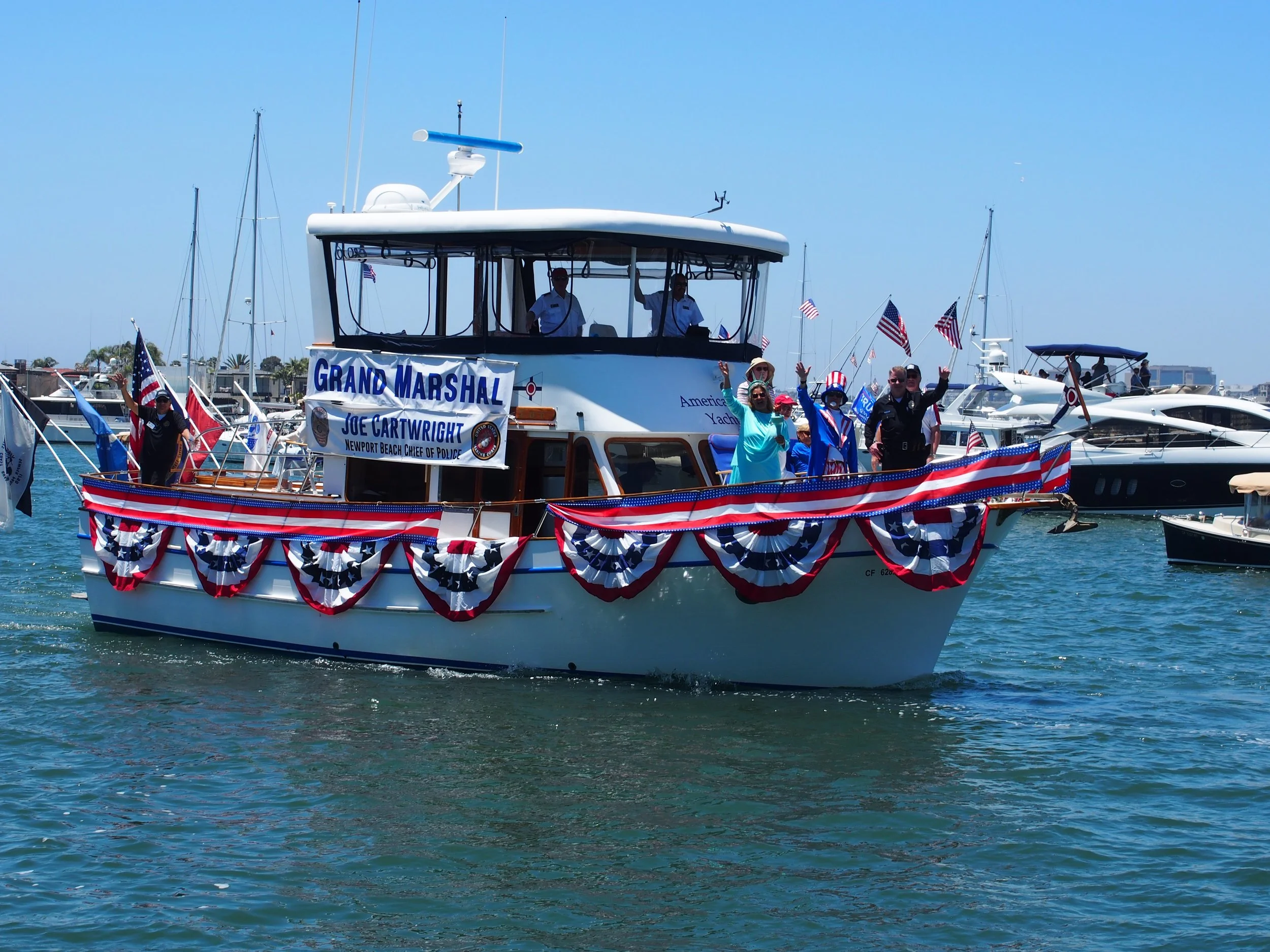 A boat decorated with red, white, and blue bunting and American flags. Several people are on the boat, waving and celebrating. The boat has a sign that reads 'Grand Marshal' and 'Joe Cartwright, Newport Beach Chief of Police.' In the background, there are other boats in a marina under a clear blue sky.