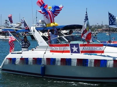 A boat decorated with American flags, patriotic banners, and a large inflatable eagle figure, celebrating America's Independence Day. The boat has a sign reading 'GOD BLESS AMERICA' and is on the water with a marina and other boats in the background.