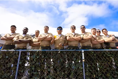 A group of ten police officers standing behind a chain-link fence with crossed arms, outdoors under a partly cloudy sky.