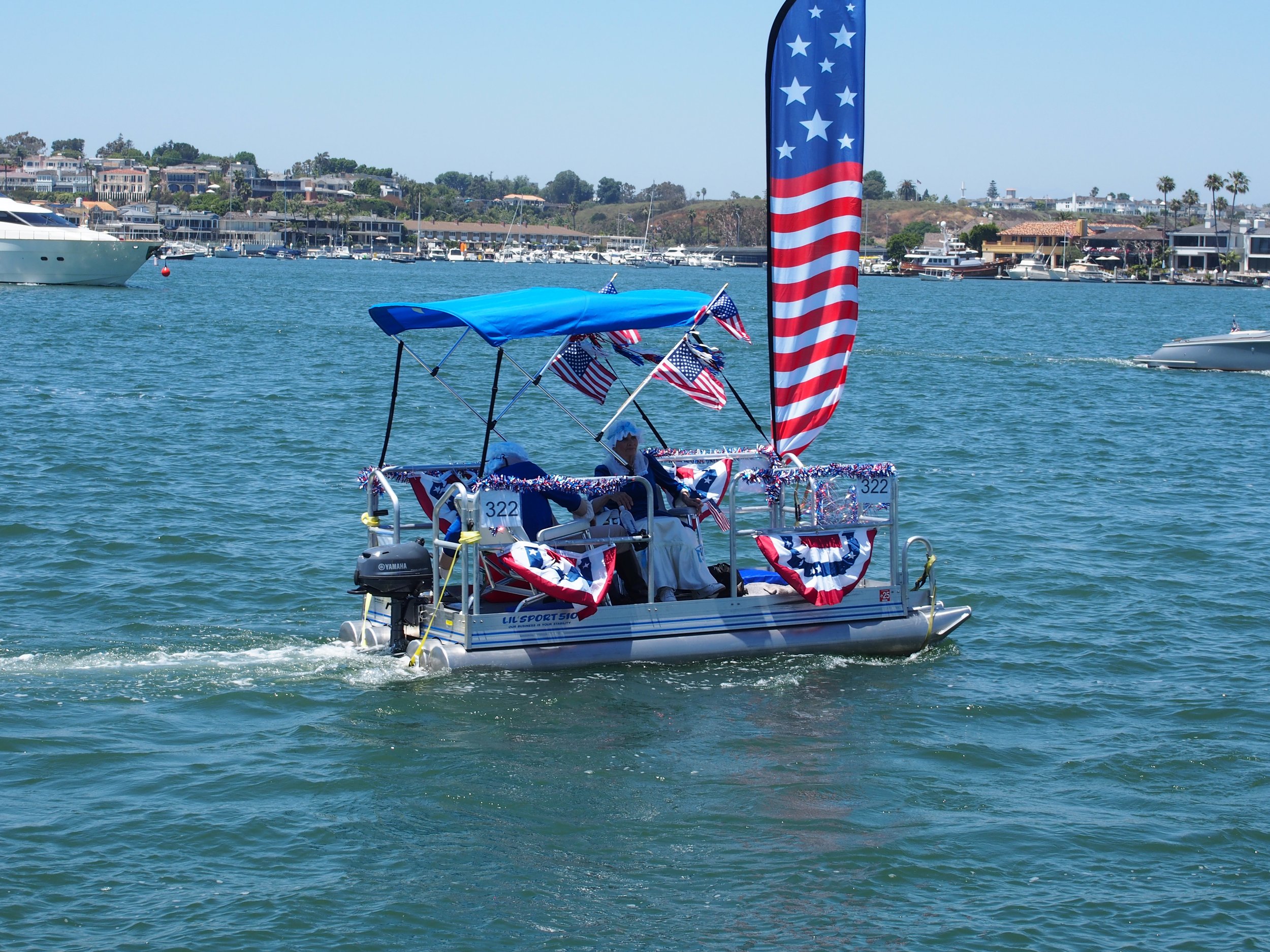 A boat decorated with American flags and red, white, and blue bunting is floating on water with a marina and hillside houses in the background.