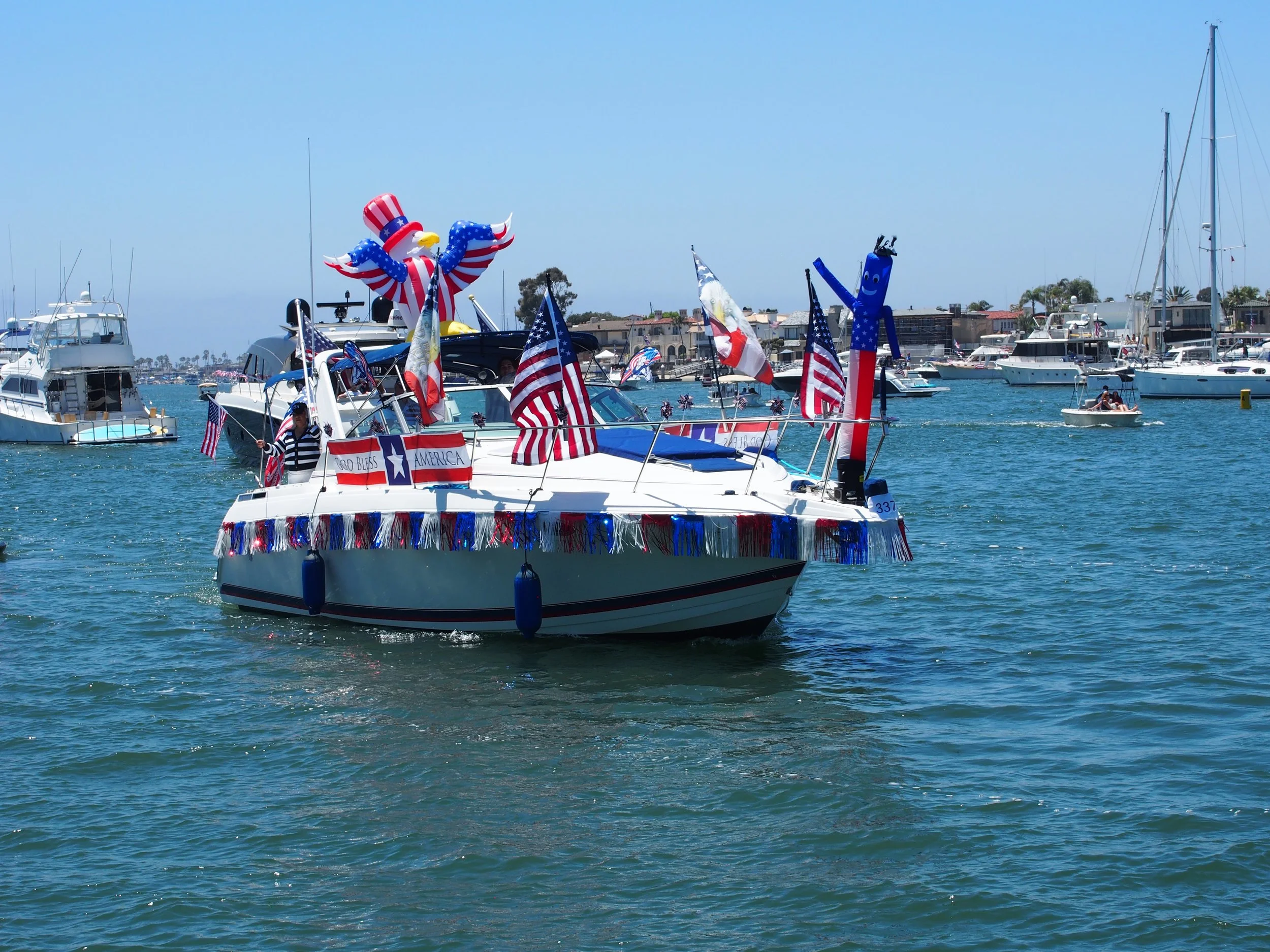 A boat decorated with American flags, red, white, and blue banners, and patriotic inflatables, including an eagle and a tall blue inflatable with a smiling face, celebrating at a marina with other boats and yachts docked nearby.