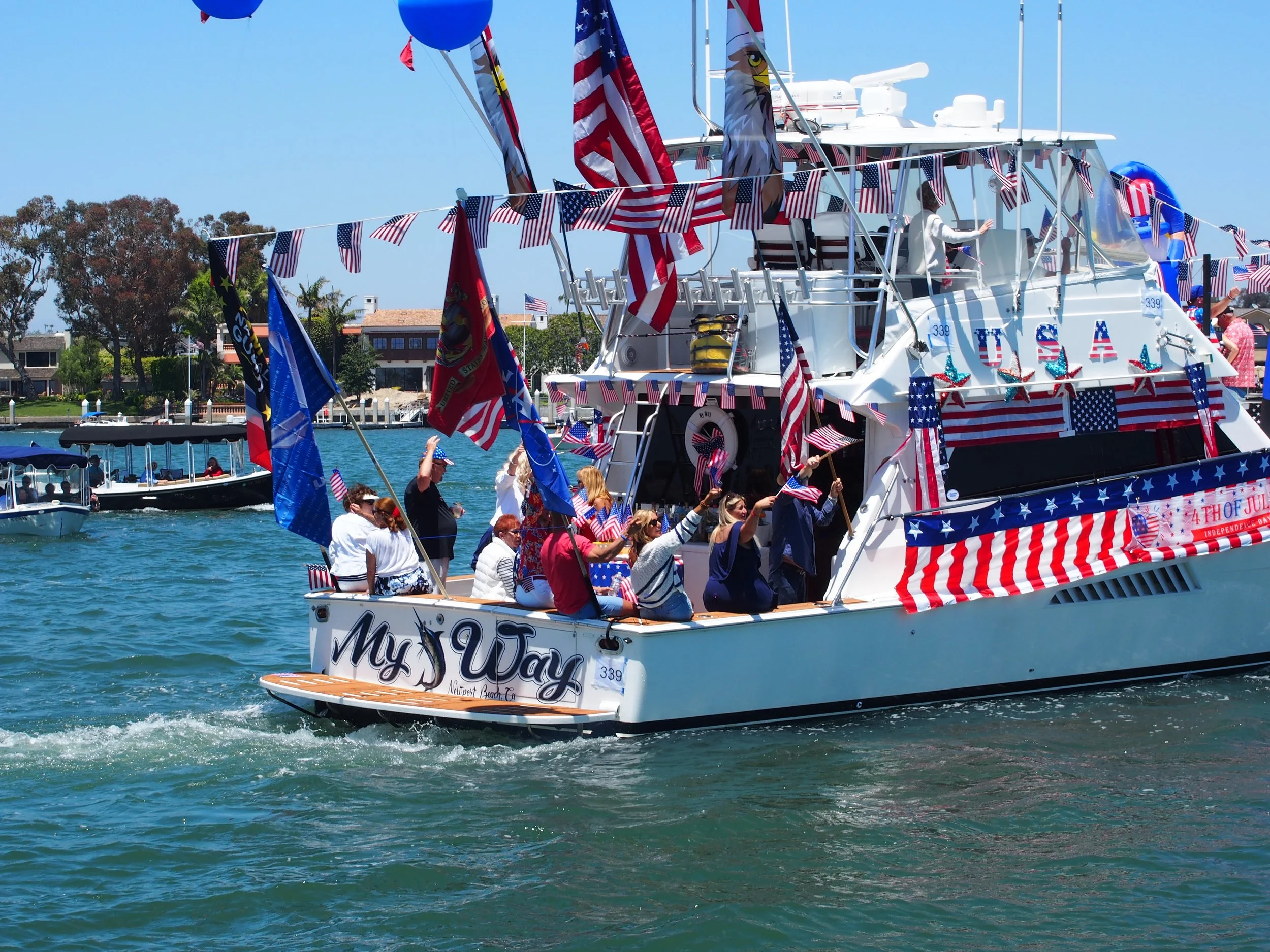A yacht decorated with American flags and patriotic items during a celebration on the water, with people waving flags and wearing patriotic clothing, tied to a dock and surrounded by other boats.