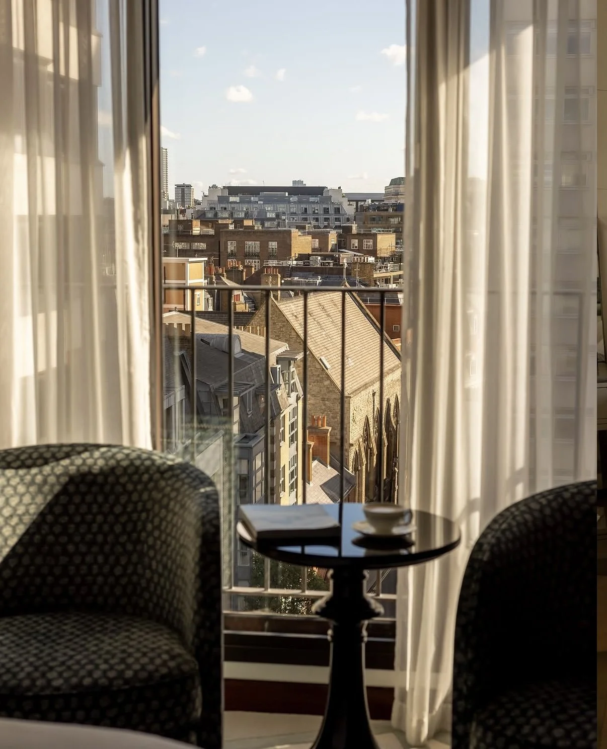 View of city skyline through window with white curtains, two chairs, and a small table with a teacup and a small bowl in a room.