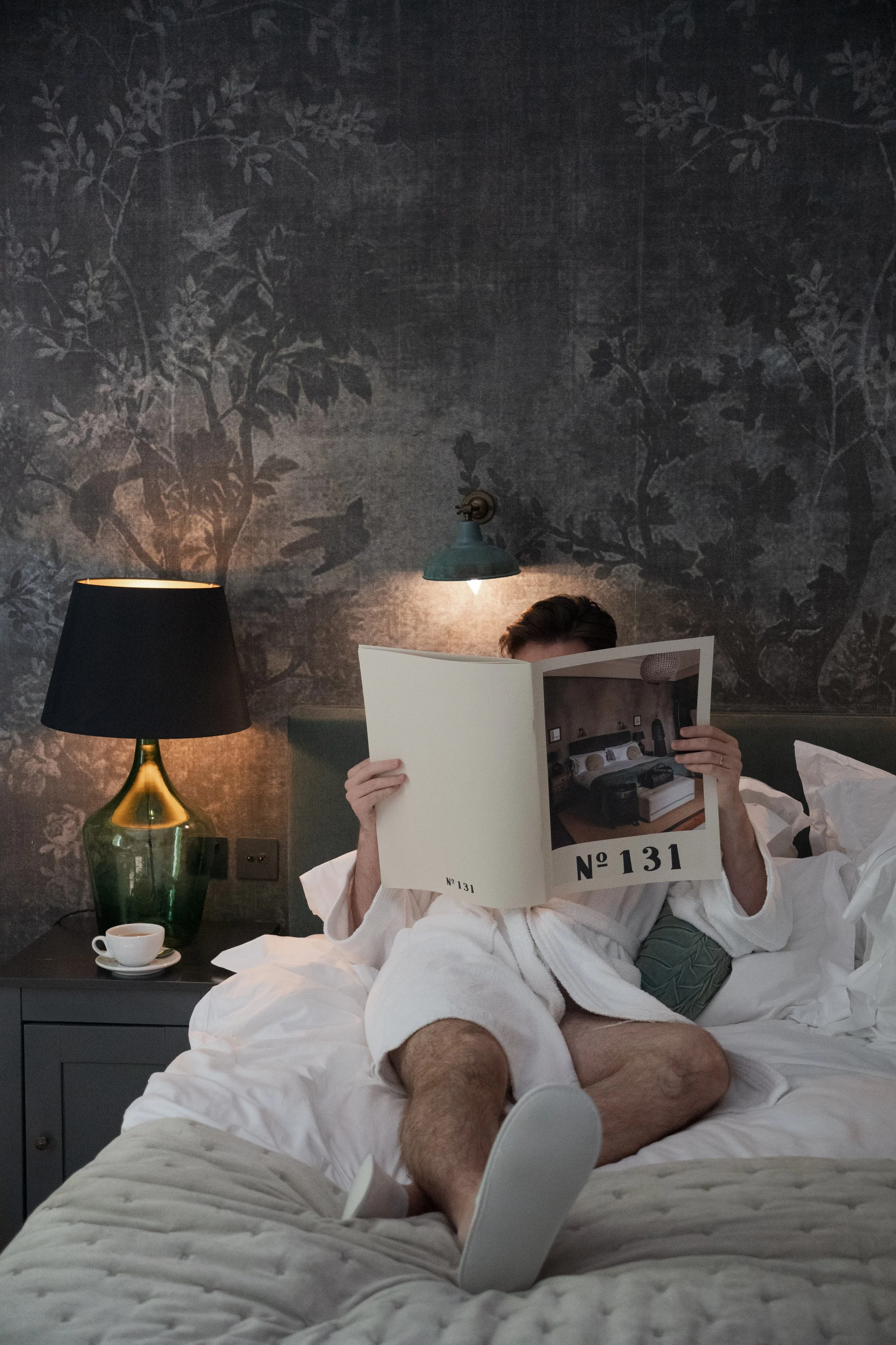 A man sitting on a bed wearing a white robe and underwear, reading a magazine labeled "No. 131." The bed has white bedding, and there's a nightstand with a lamp, a cup, and other items next to him. The background features a dark wall with a floral pattern, a lampshade, and a wall-mounted light fixture.