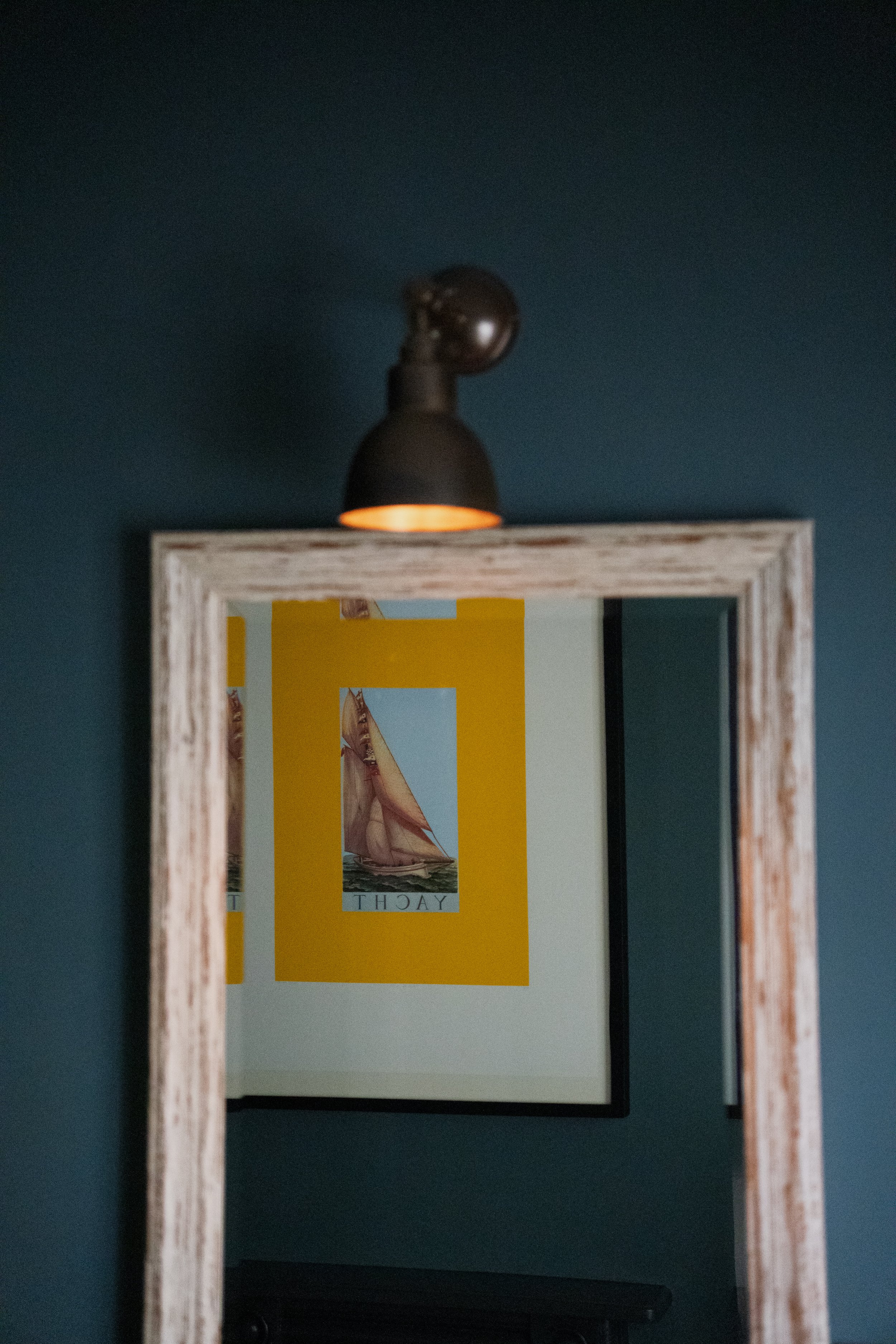 A wooden-framed mirror on a blue wall reflects a poster with a yellow background, an illustration of a sailing ship, and the word 'YACHT' at the bottom.