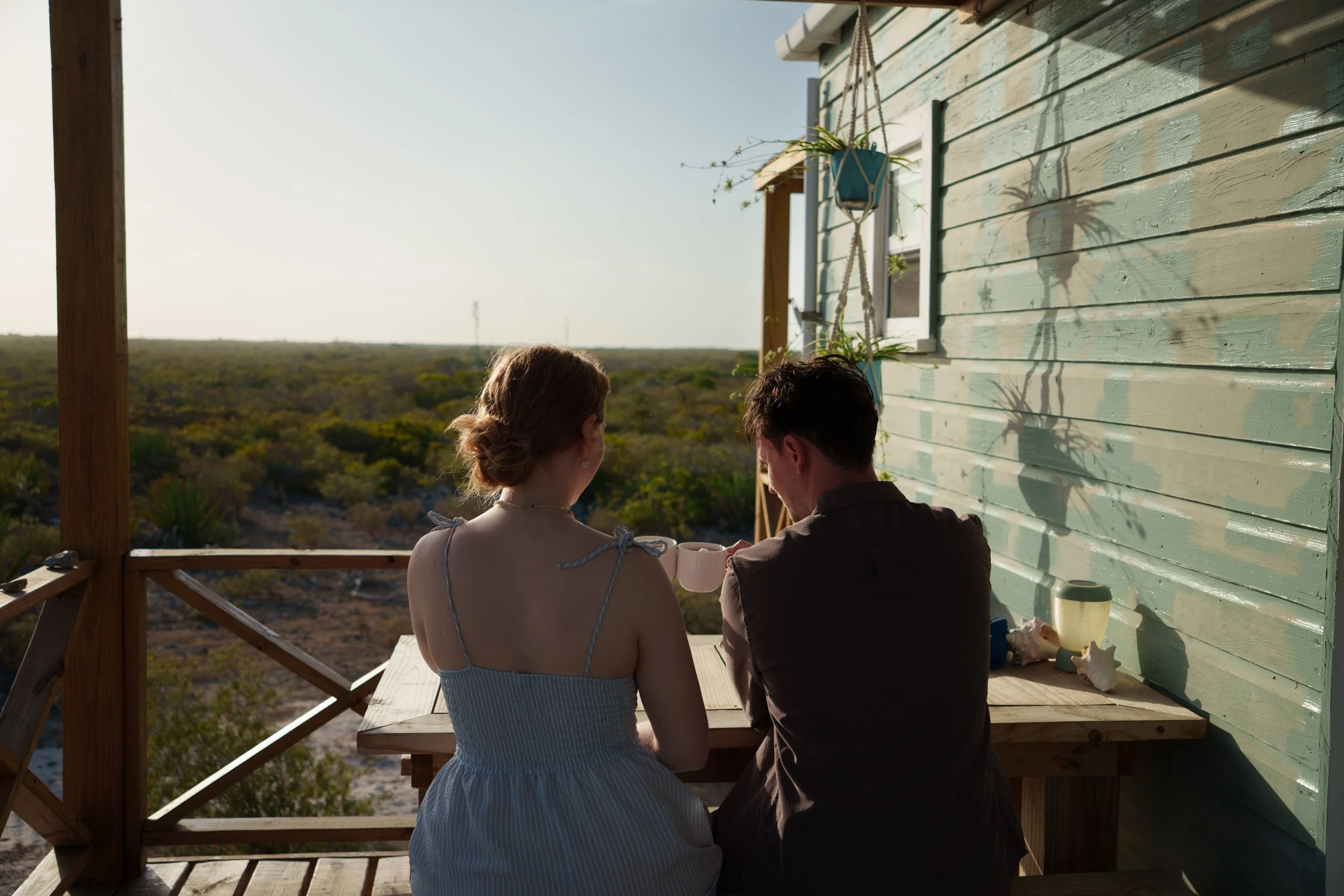 Two people sitting at a wooden table on a balcony, facing a scenic landscape. The woman has her hair in a bun and wears a sleeveless dress, while the man has dark hair and is dressed in a dark shirt. The sunlight casts shadows of plants on the green wooden wall.