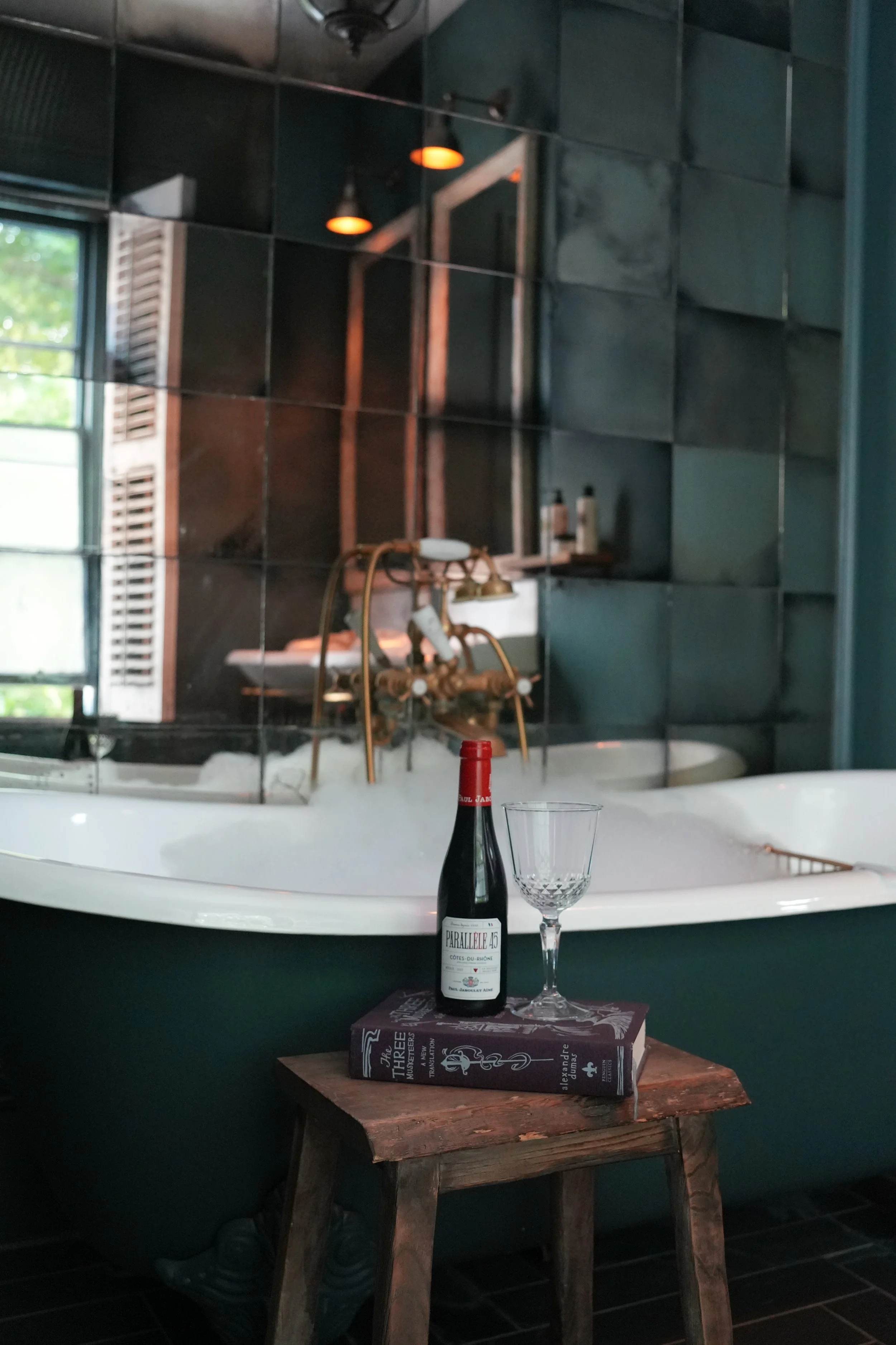 A stool with a bottle of red wine, a wine glass, and a book in front of a mirror that reflects a bathroom with green tiled walls, a rolltop bathtub, and a brass faucet. There is a window with shutters and some bottles on a shelf in the background.