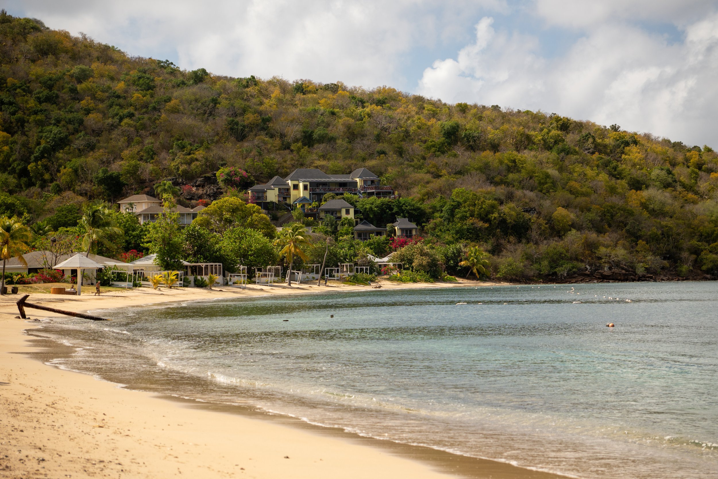 A Caribbean beach with light coloured sand, calm water, and a backdrop of houses on a hillside covered with green and brown trees under a partly cloudy sky. A large anchor rests on the sand.