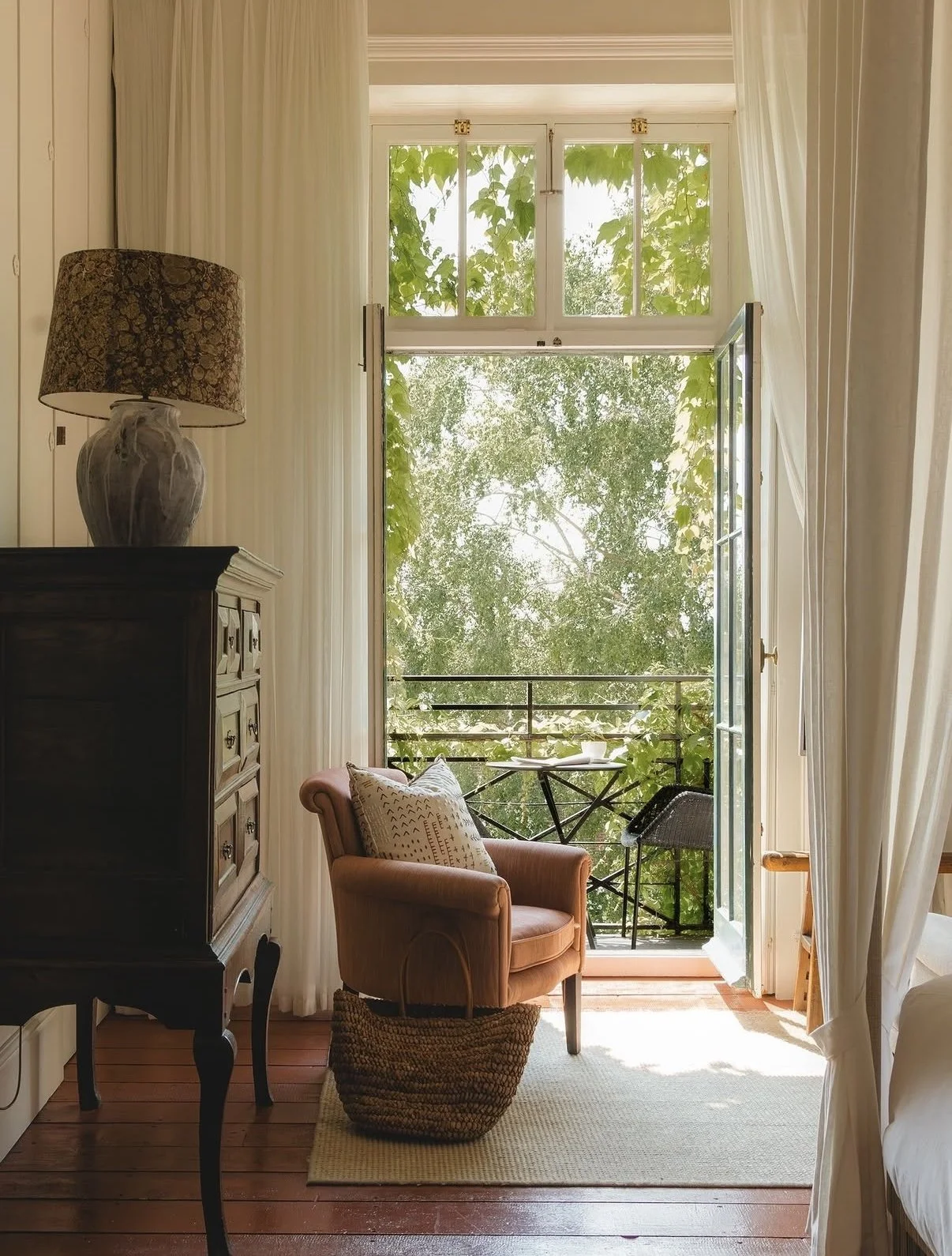 A cozy room with an open door leading to a small balcony with trees outside. Inside, there's a pink armchair with a decorative pillow, a woven basket in front, an ornate wooden dresser with a lamp, and white curtains framing the doorway.