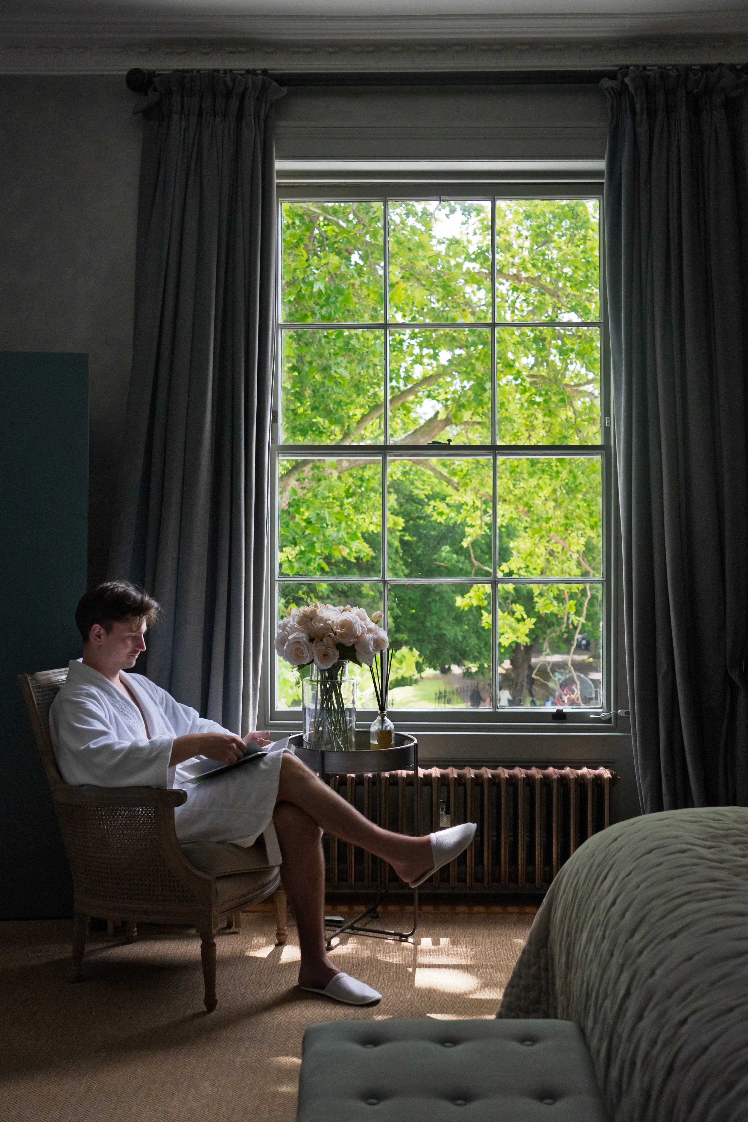 A man sitting in a wooden chair by a window, reading a book. The room has dark curtains, a vase with white roses on a side table, and a bed with a greenish comforter in the foreground.