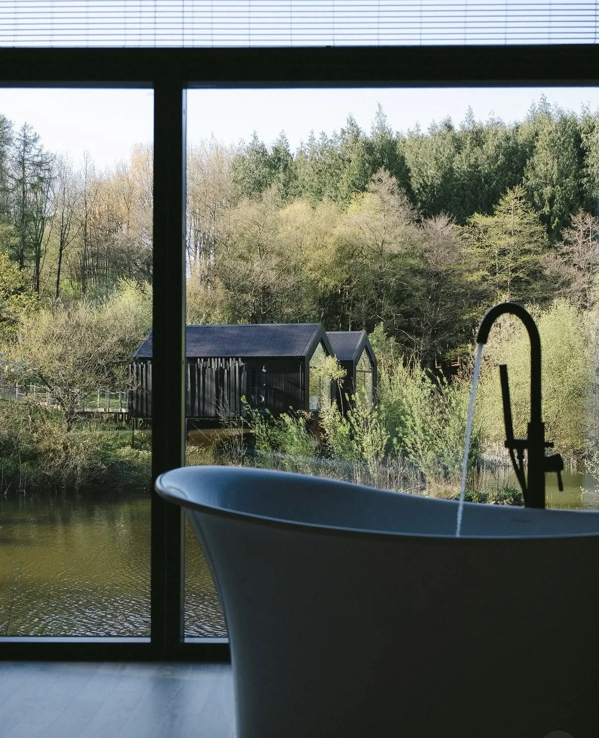 View through a large window featuring a bath sink with a black faucet, overlooking a pond, grassy landscape, trees, and black wooden buildings in the distance.
