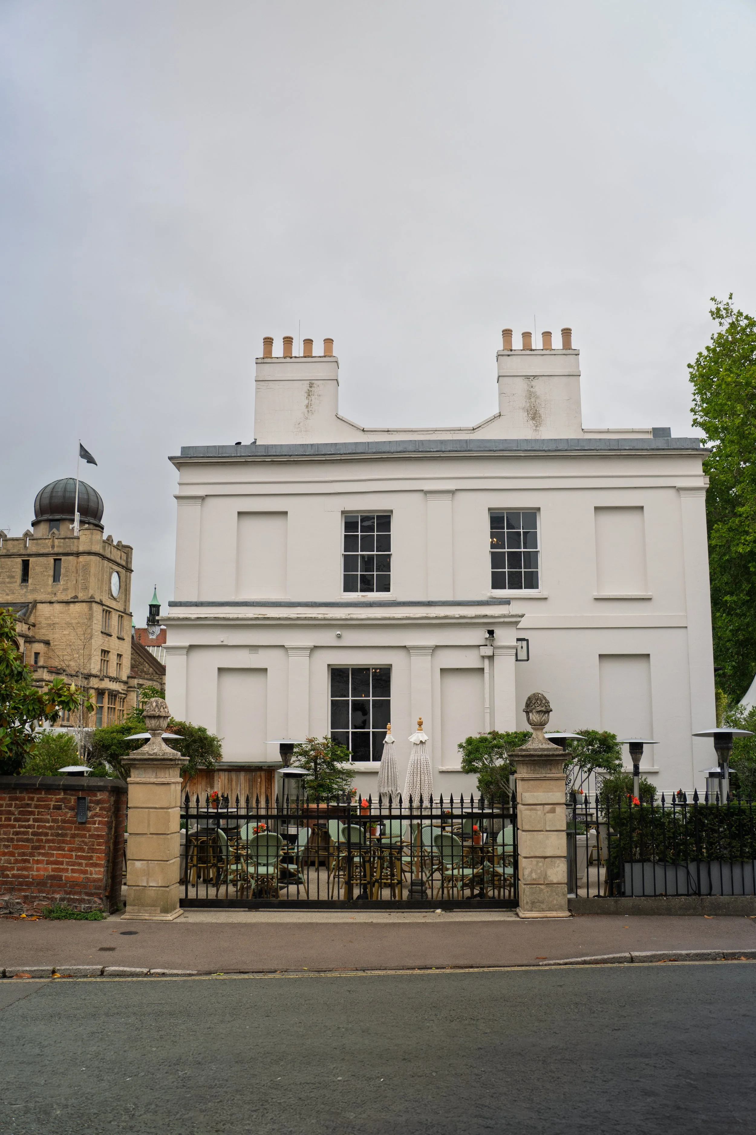 White historic building with multiple windows, surrounded by a small courtyard with garden furniture, parasols, and a black wrought-iron fence, under a cloudy sky.