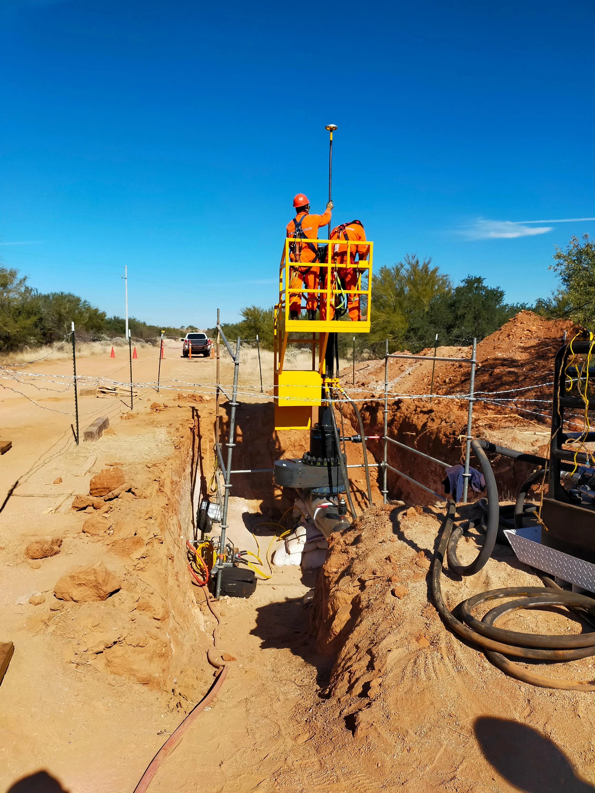 Workers in orange safety gear and helmets operating equipment in a construction or utility work site on a clear, sunny day with a blue sky and scattered clouds.