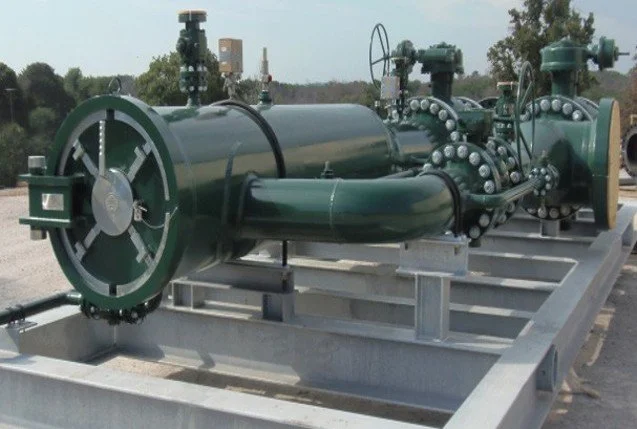 Large industrial pressure vessel or pump system on a metal platform outdoors with trees and sky in the background.