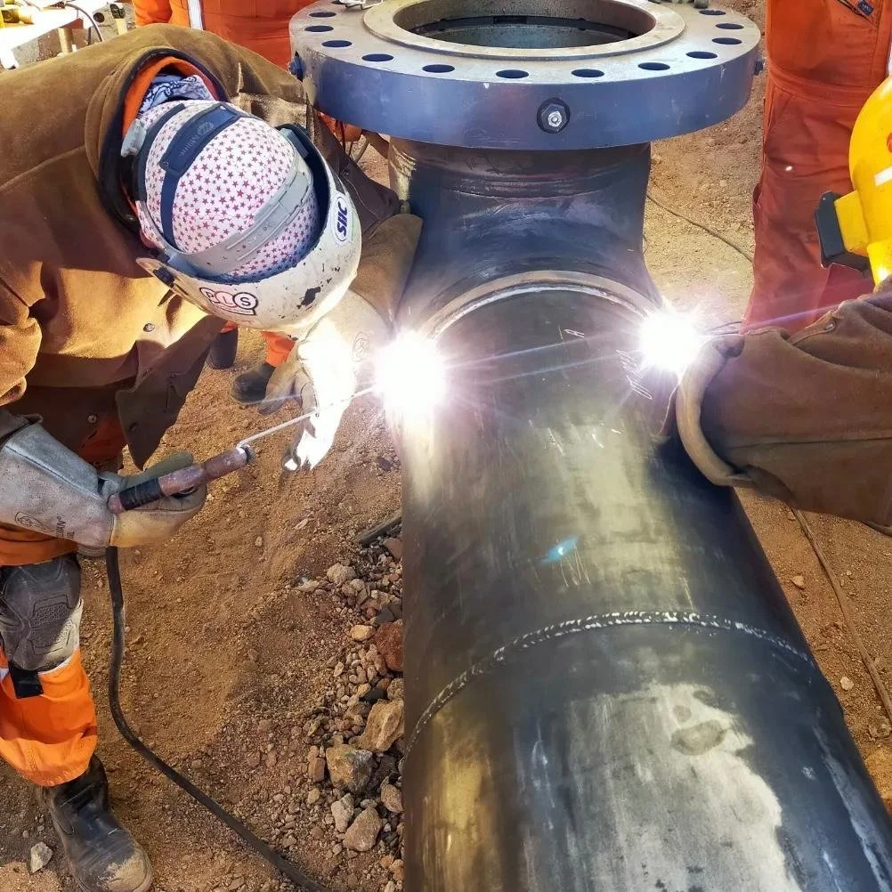 Two workers are welding a large metal pipe, wearing protective helmets and gloves, with bright sparks and light from the welding process.