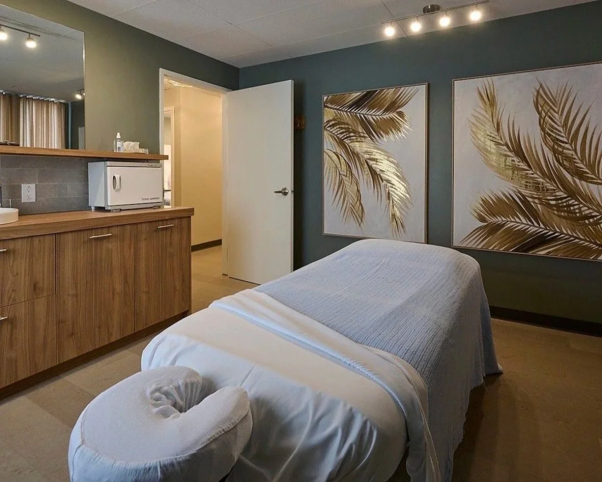 A massage room with a massage table covered with a white sheet and a gray blanket. Two large framed artworks of palm leaves hang on the teal wall behind the table. There's a wooden cabinet with a microwave and a tissue box, and an open door leads to a hallway.