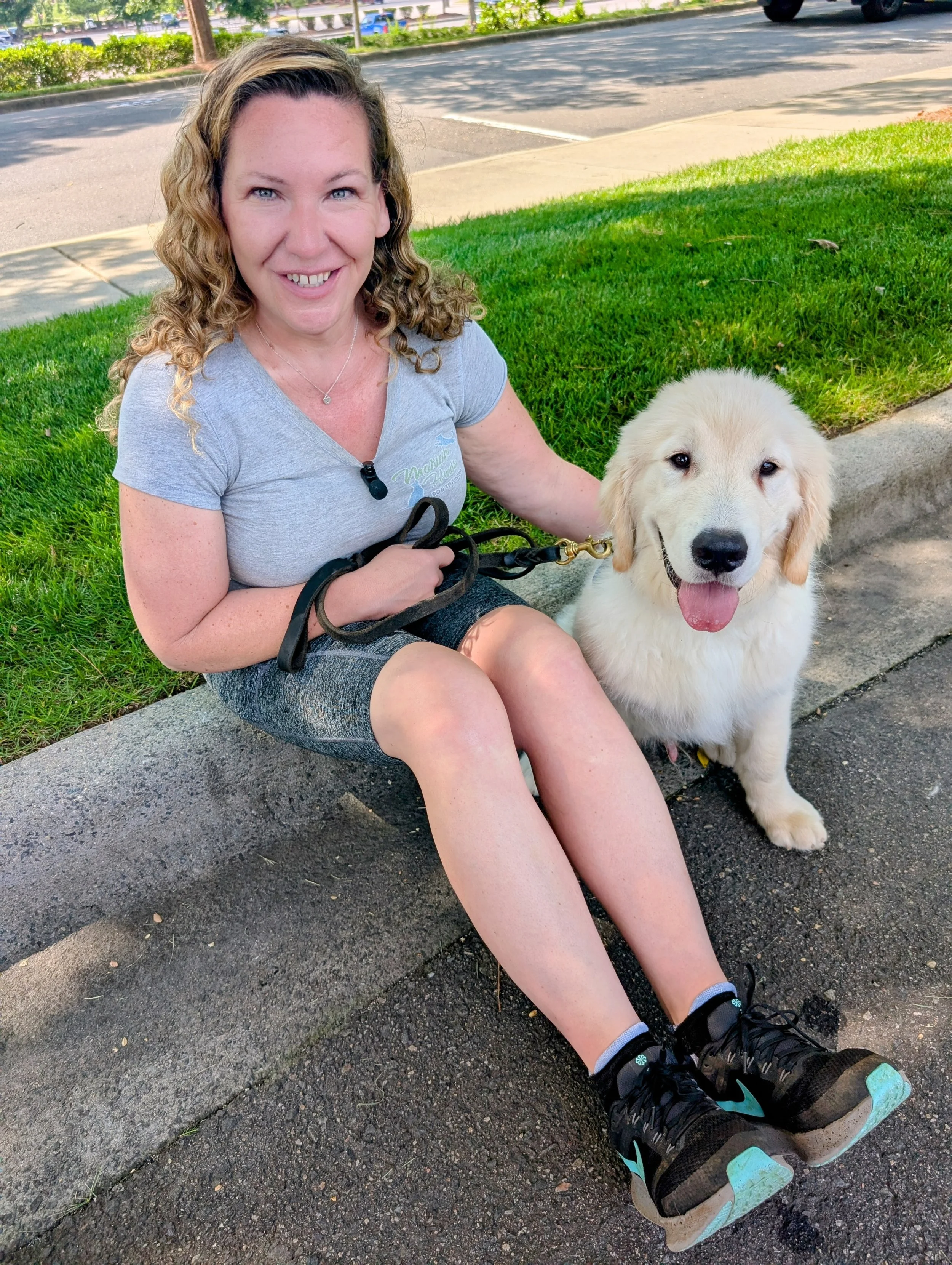 A woman with curly blonde hair sitting on a curb on a sidewalk, smiling, holding a leash attached to a happy golden retriever puppy, with a parking lot and trees in the background.