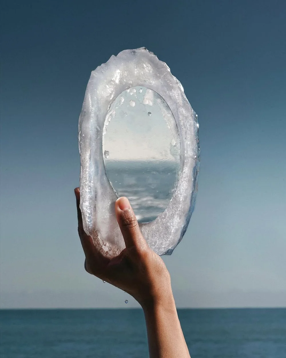A hand holding up a mirror made of ice against the sea