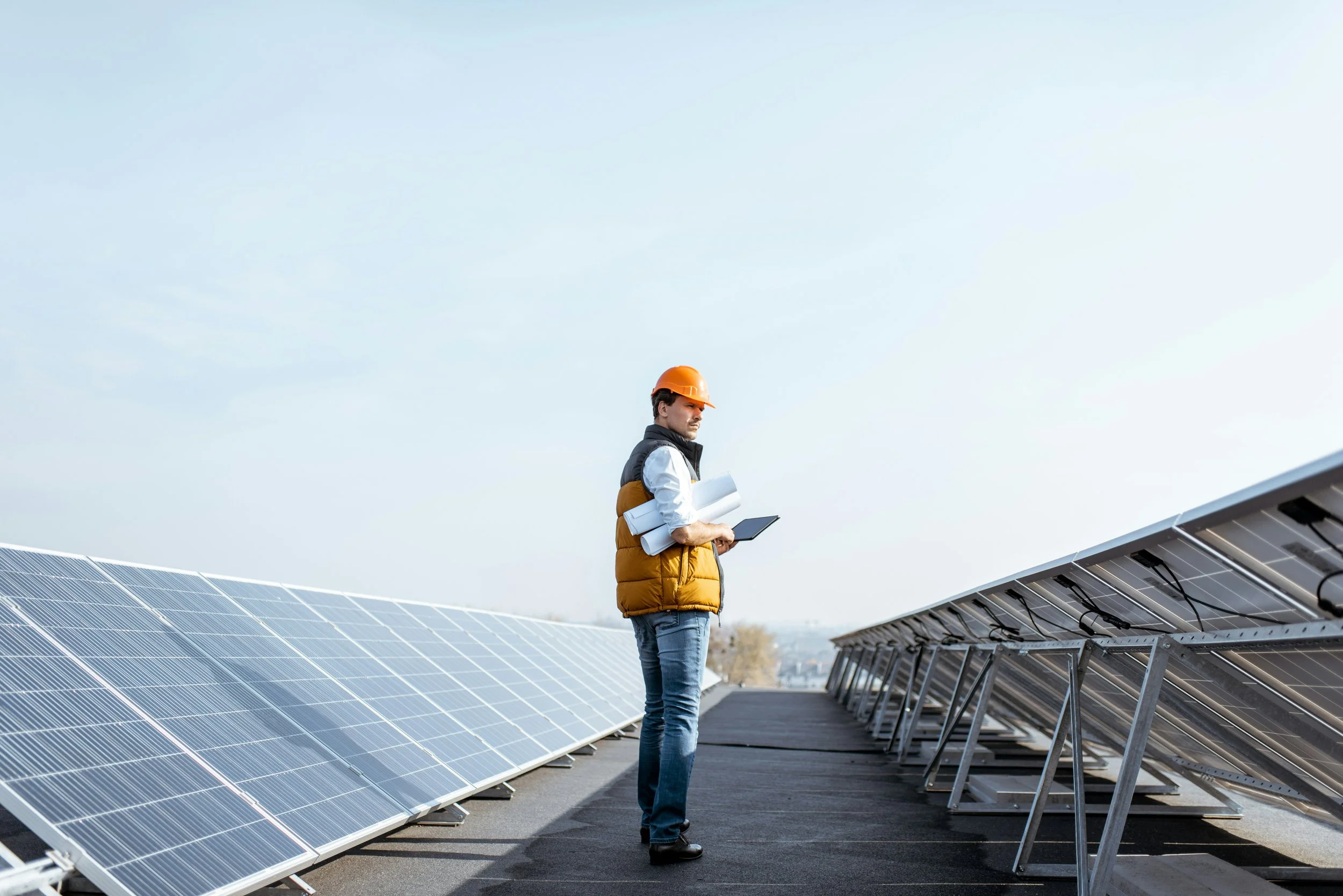 A man wearing an orange hard hat and a yellow vest inspecting solar panels with a tablet and rolled-up plans, standing between rows of solar panels outdoors under a clear sky.