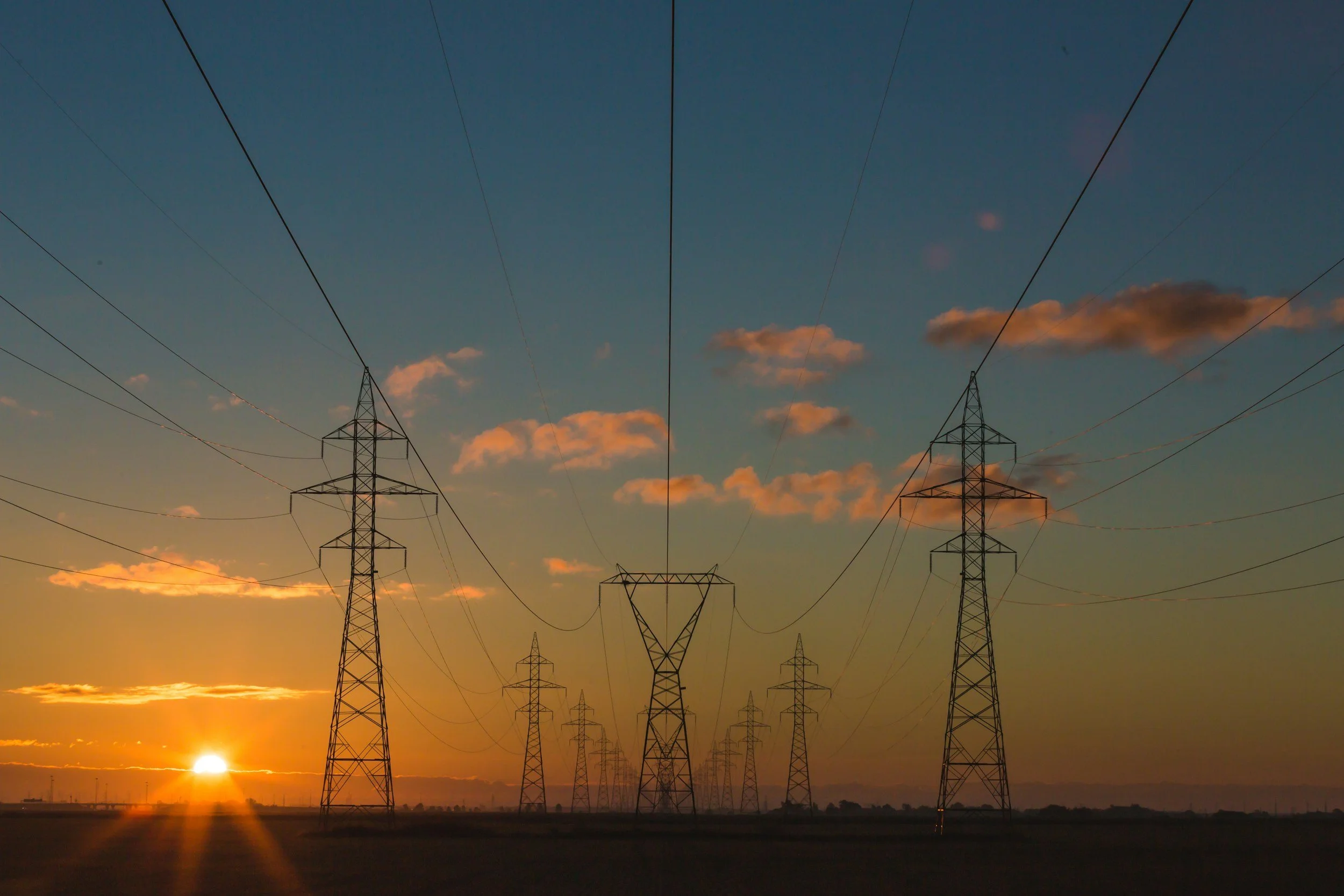 A landscape with high-voltage power lines and towers at sunset, with a partly cloudy sky and the sun near the horizon.