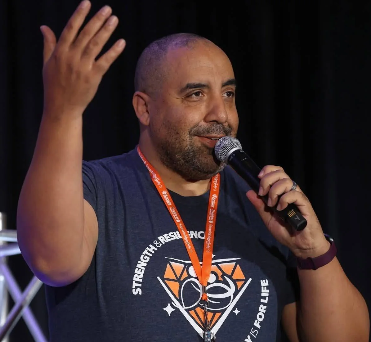 A man speaking into a microphone at an event, wearing a dark shirt with a diamond and fist graphic and a lanyard around his neck.