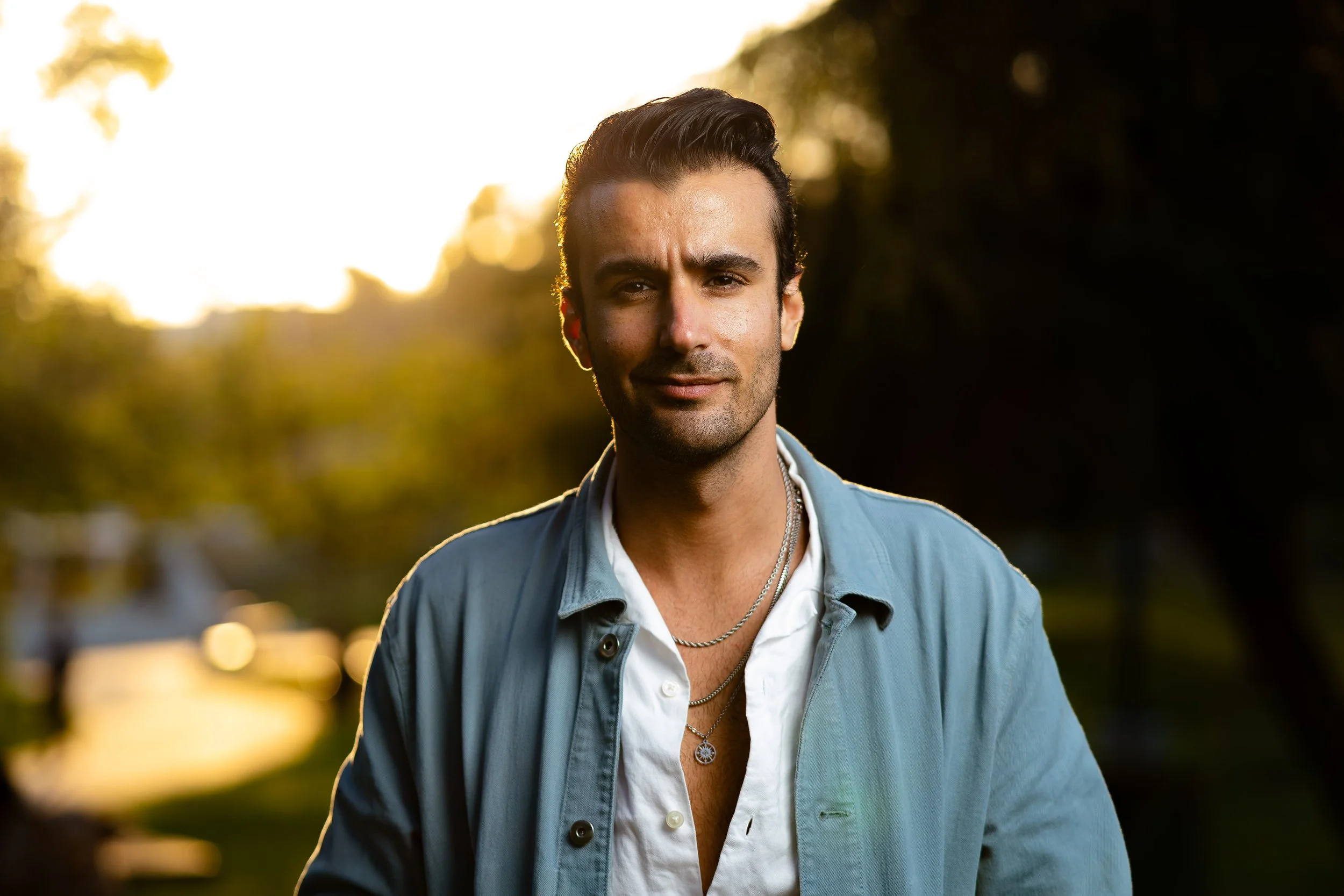 A young man with dark, slicked-back hair and light skin looking directly at the camera. He is wearing a white shirt underneath a blue jacket with the top few buttons open, revealing layered necklaces. The background features blurred trees and the warm glow of sunlight, suggesting late afternoon or early evening.