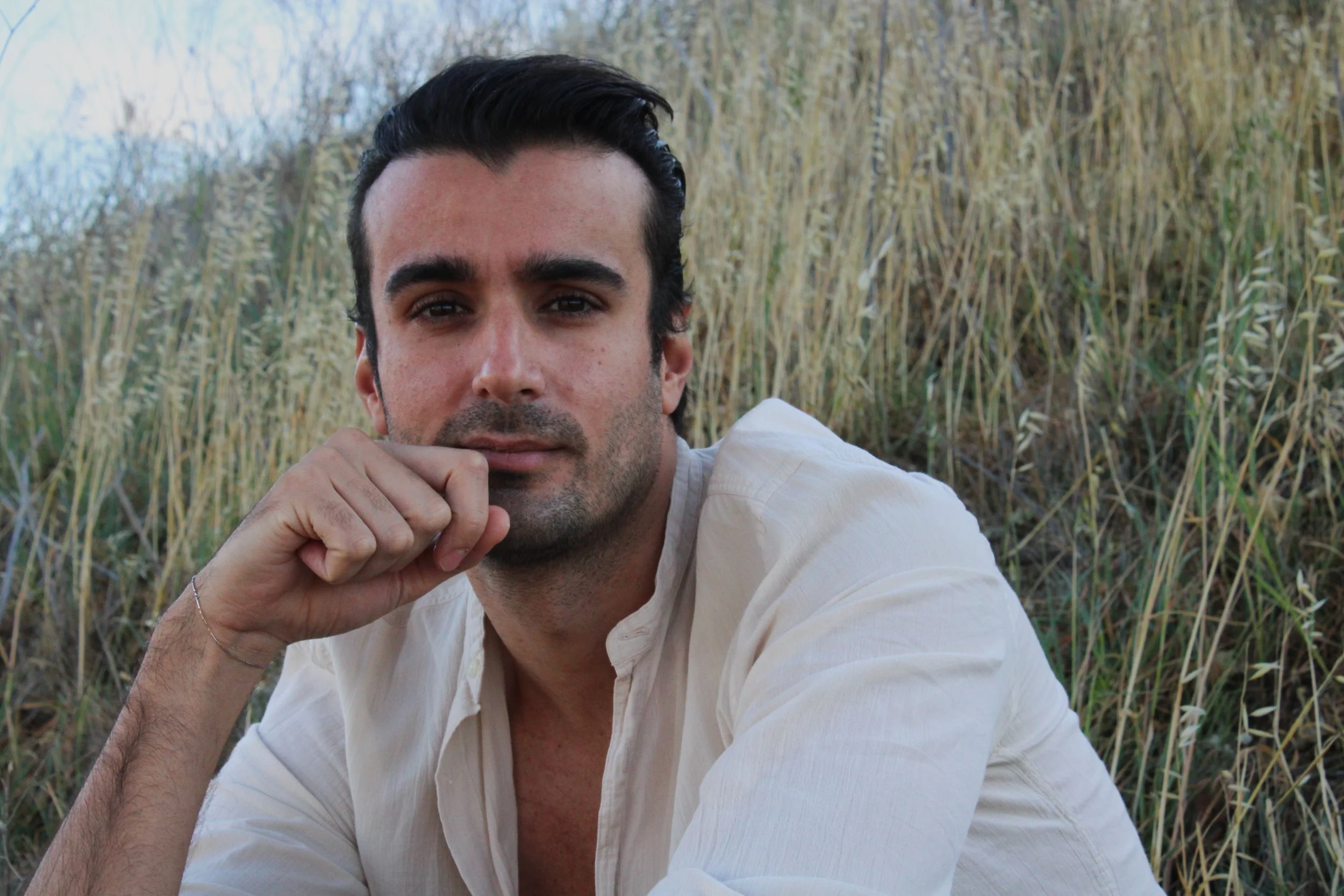 A young man with dark hair and light skin lying on the ground in front of tall grasses, resting his chin on his hand and looking into the camera.