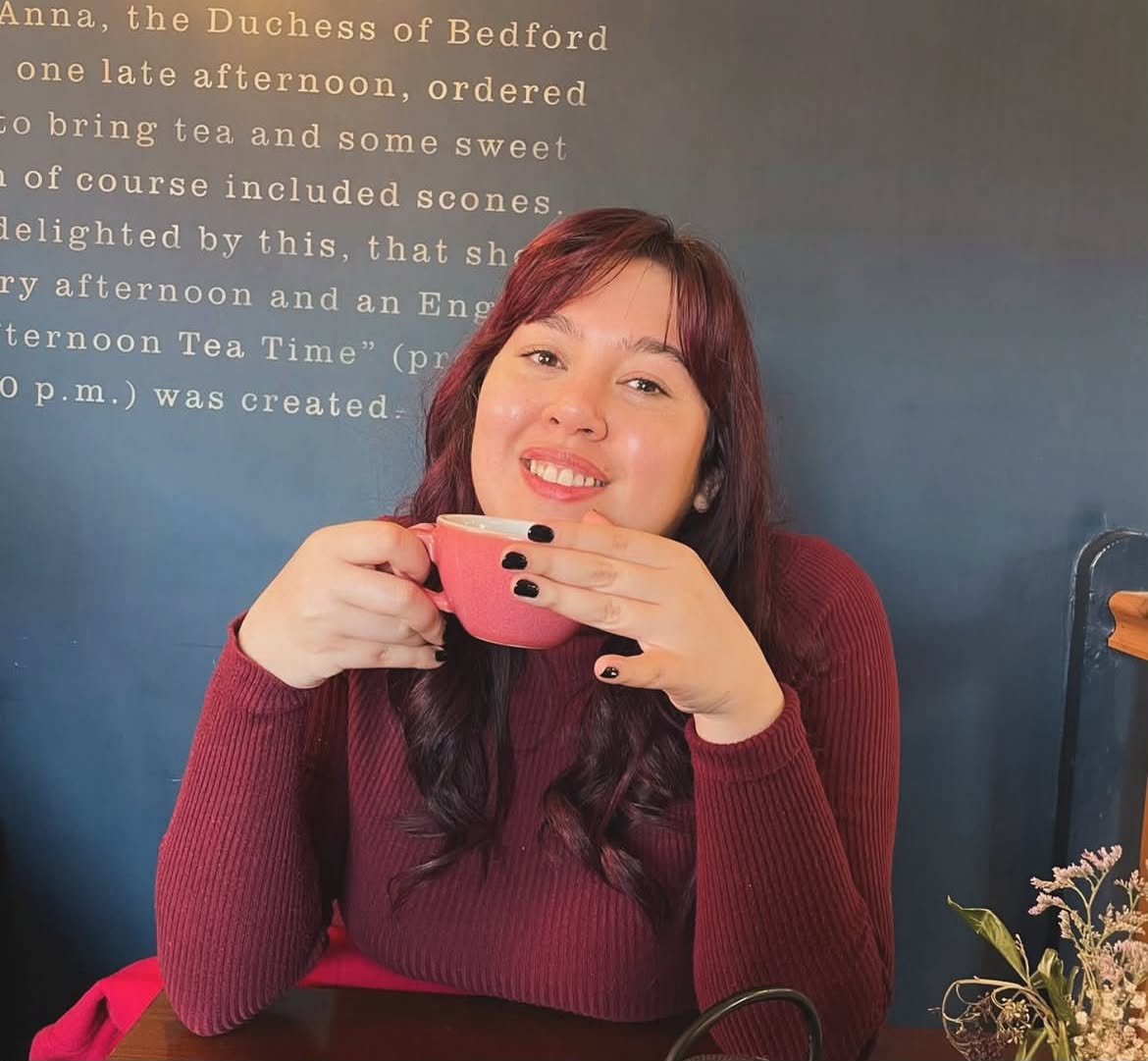 A woman with long dark hair and light skin, smiling while holding a pink coffee cup, sitting at a table in a café with a dark background and text on the wall.