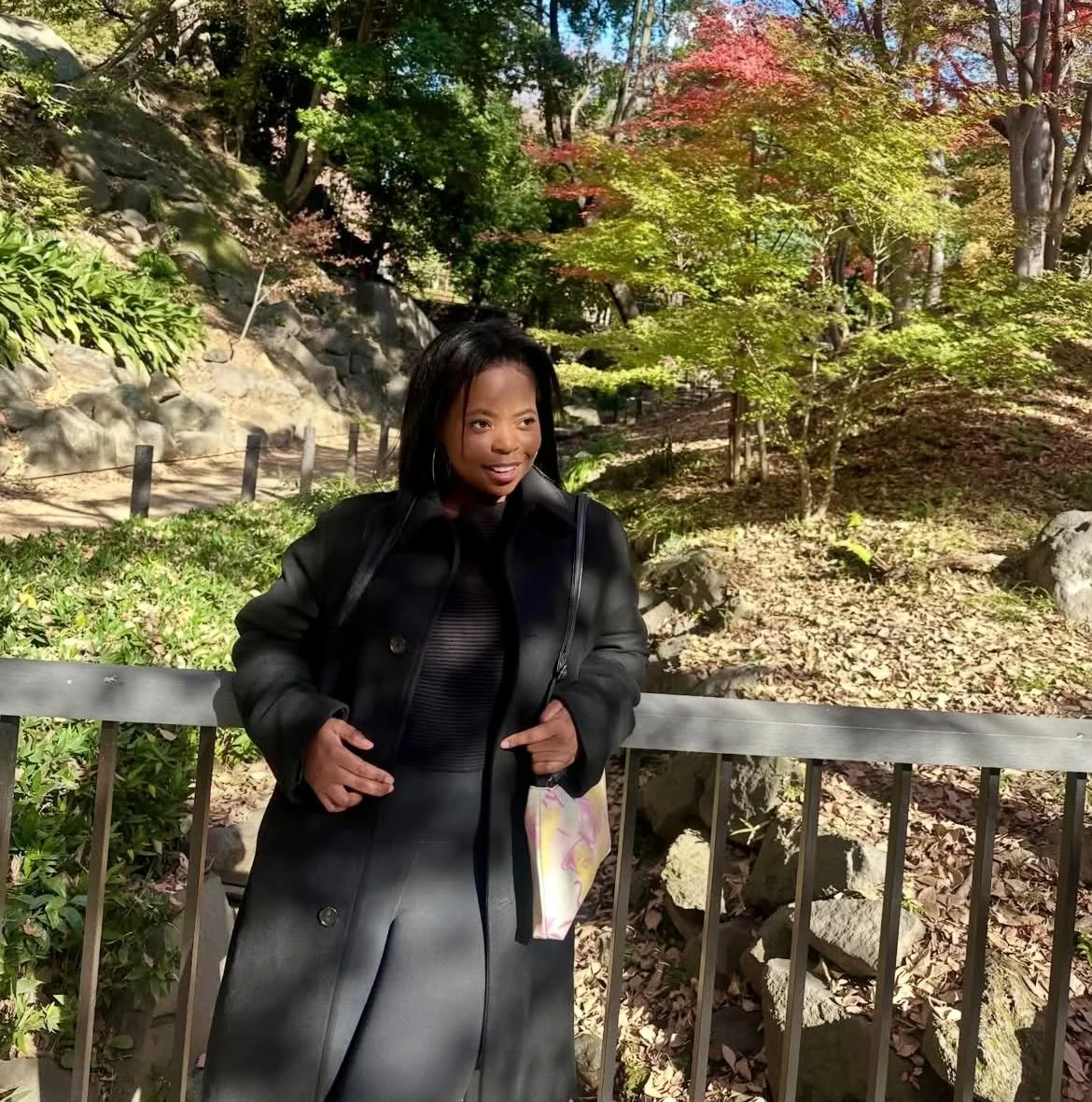 A woman in a black coat standing by a railing in a park with trees and rocks, autumn foliage, and sunlight.