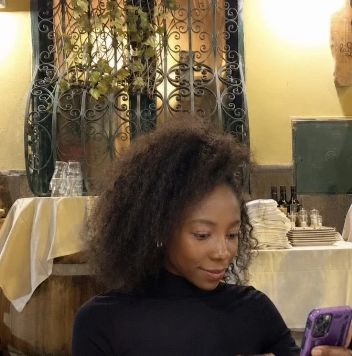 A woman with curly hair looking at her phone in a restaurant with yellow walls, a decorative iron window grill, and a table set with glasses, dishes, and bottles.