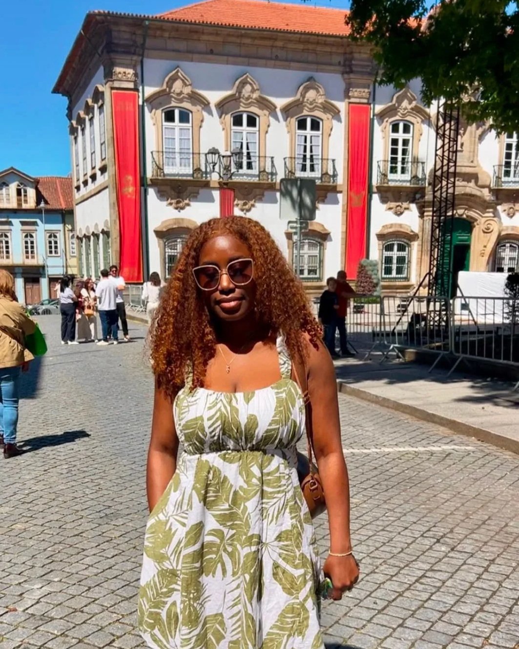 A woman with brown curly hair wearing a tropical patterned dress, sunglasses, and a bracelet standing on cobblestone street in front of a decorative building with red banners and a small crowd in the background.