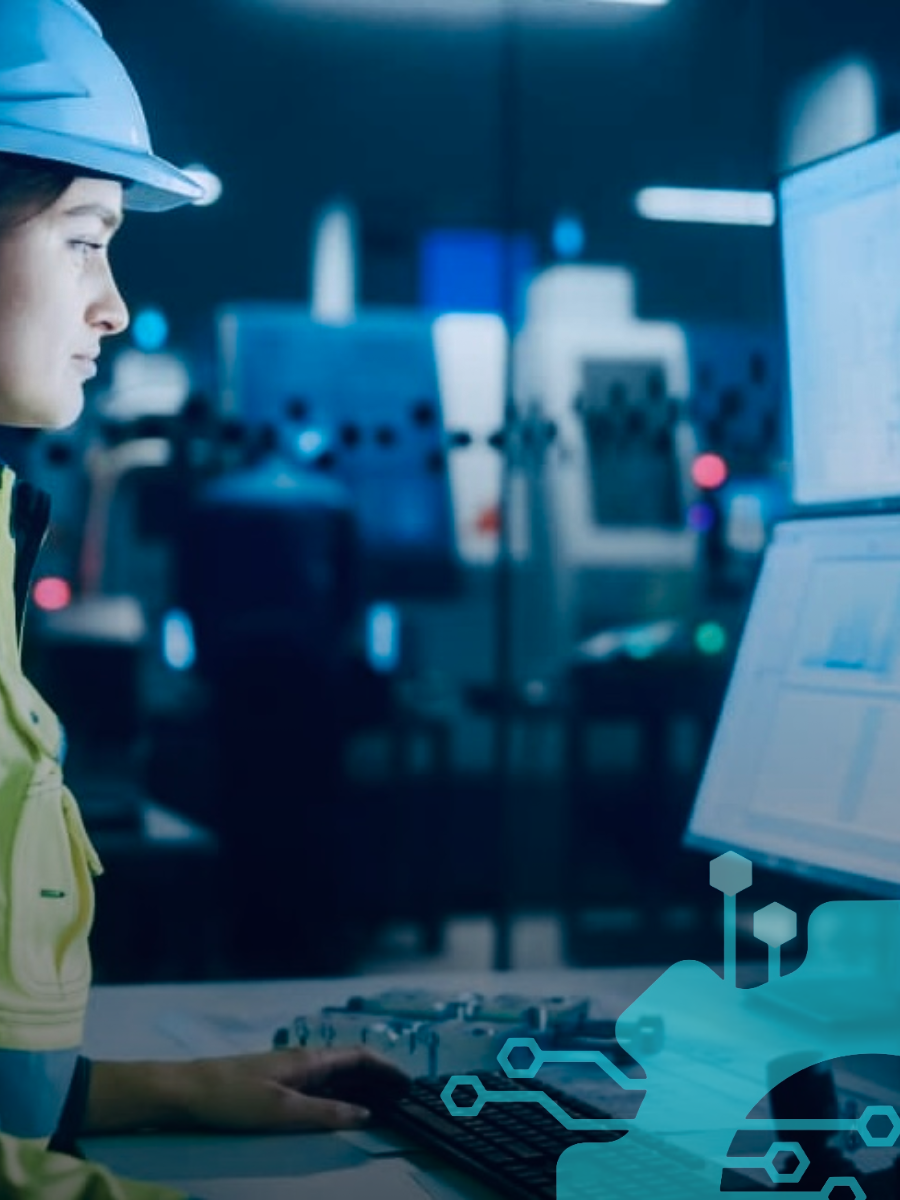 A female engineer in a hard hat working at a computer in an industrial or manufacturing setting with machinery in the background.