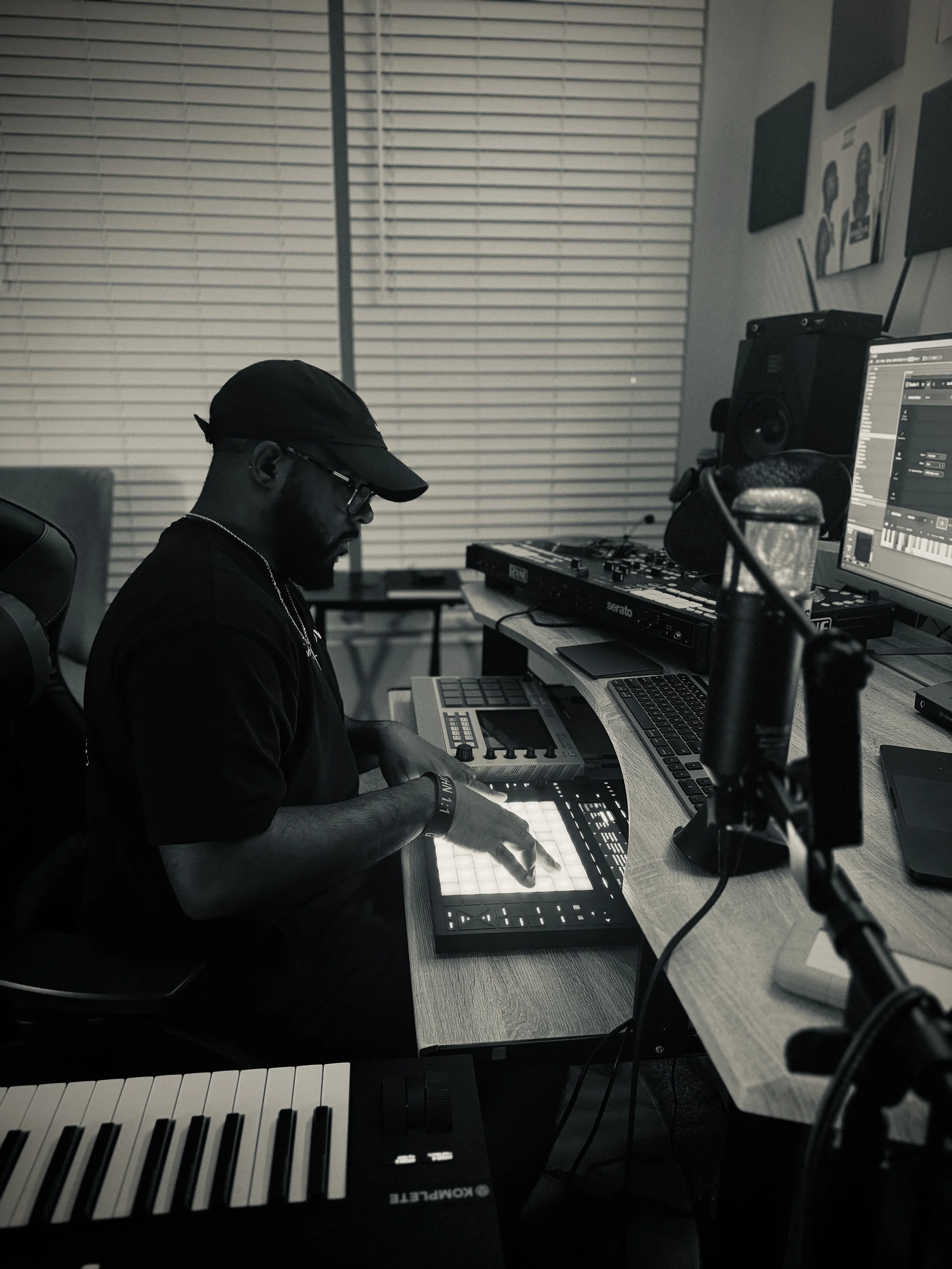 A person wearing a black cap and glasses working on music production in a home studio. They are using a grid-based MIDI controller with illuminated pads, surrounded by a keyboard, computer monitor, speakers, and other music equipment.