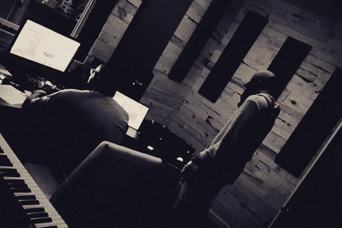 Two people in a music studio; one person sitting at a computer and the other standing near a keyboard, with wood-paneled walls.