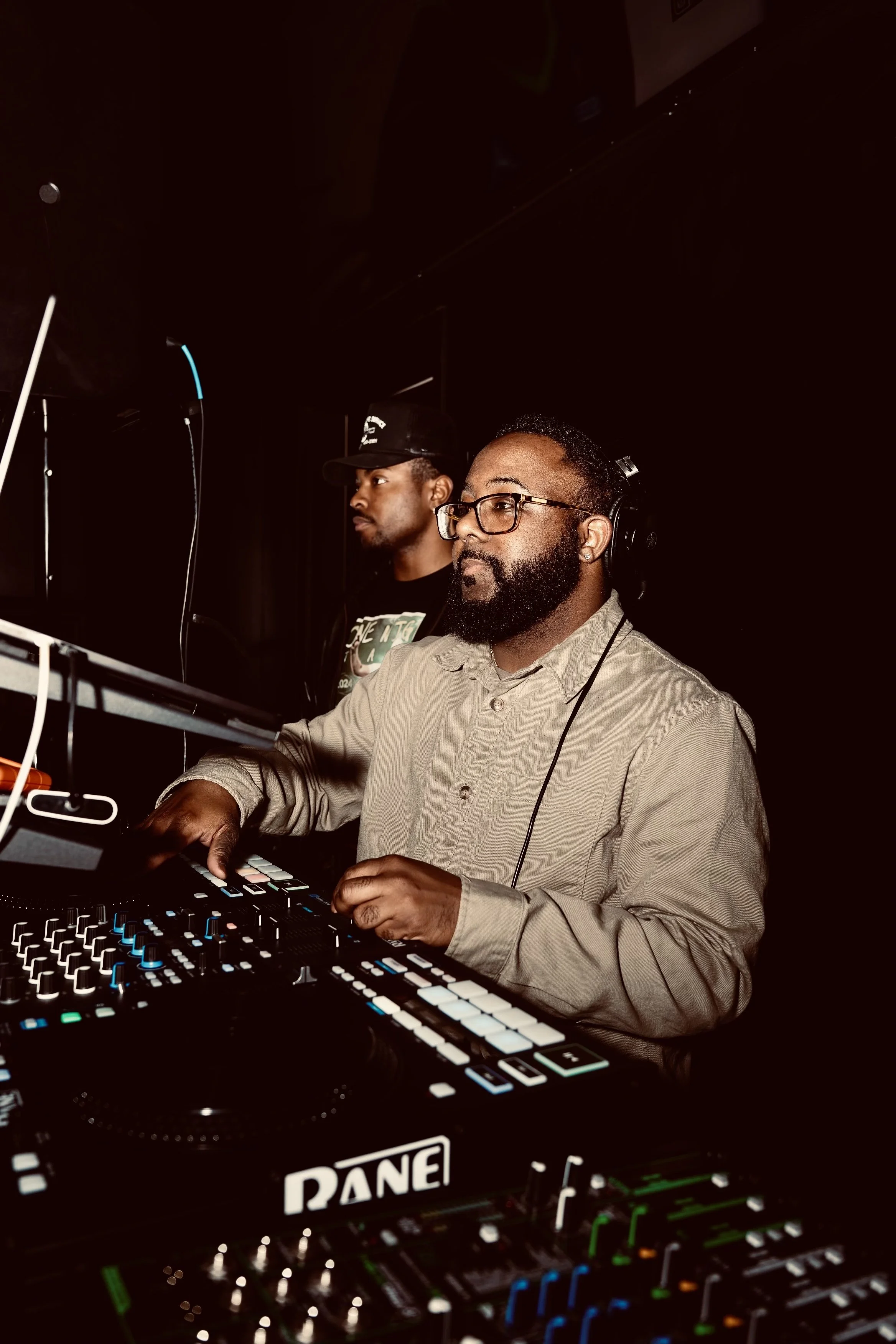 Two men operating DJ equipment in a dimly lit setting. One man is wearing headphones, glasses, and a beige shirt, focused on the controls, while the other man is in the background wearing a black cap and t-shirt.