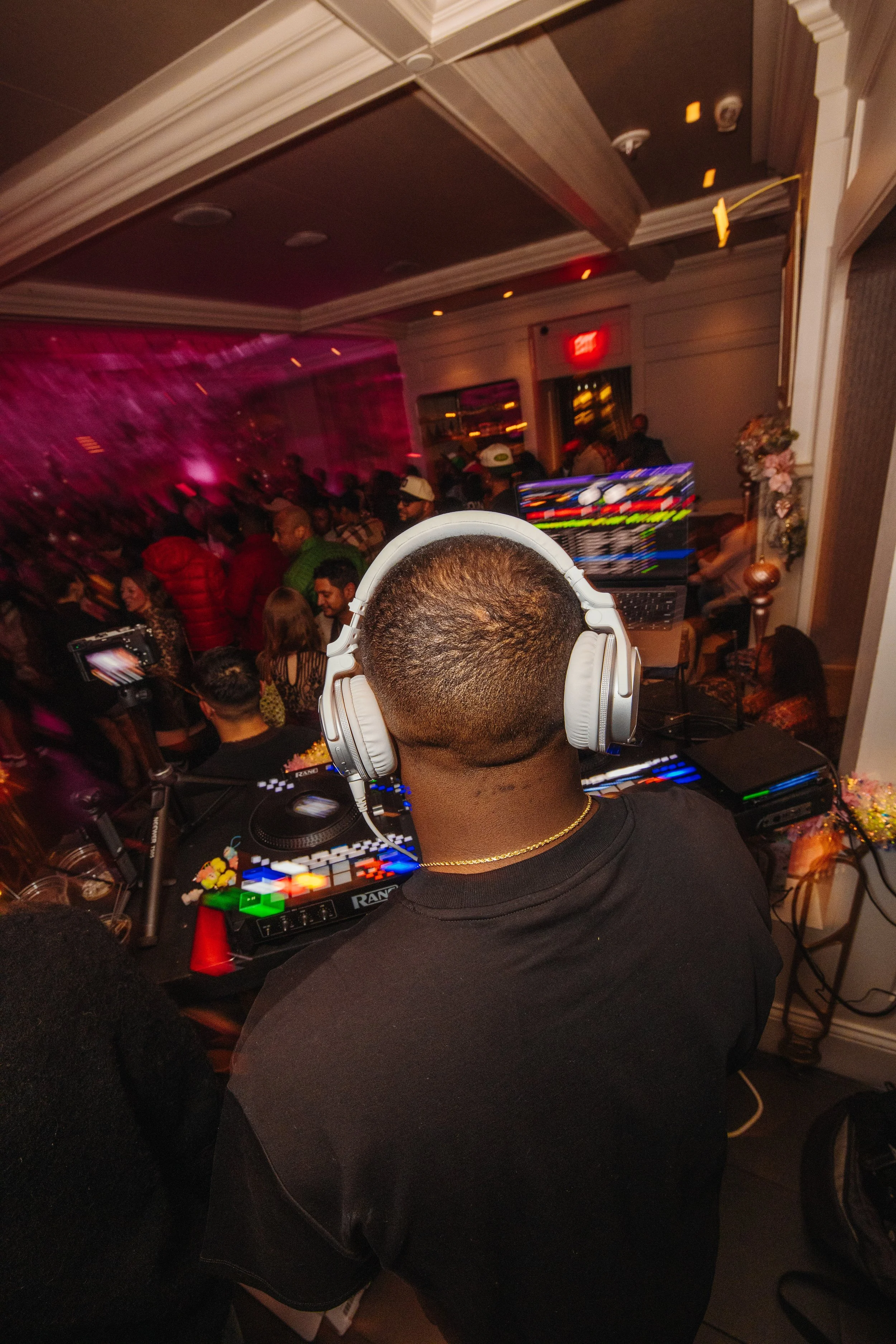 A DJ wearing large headphones is playing music at a crowded party or event, with people dancing and socializing in the background.