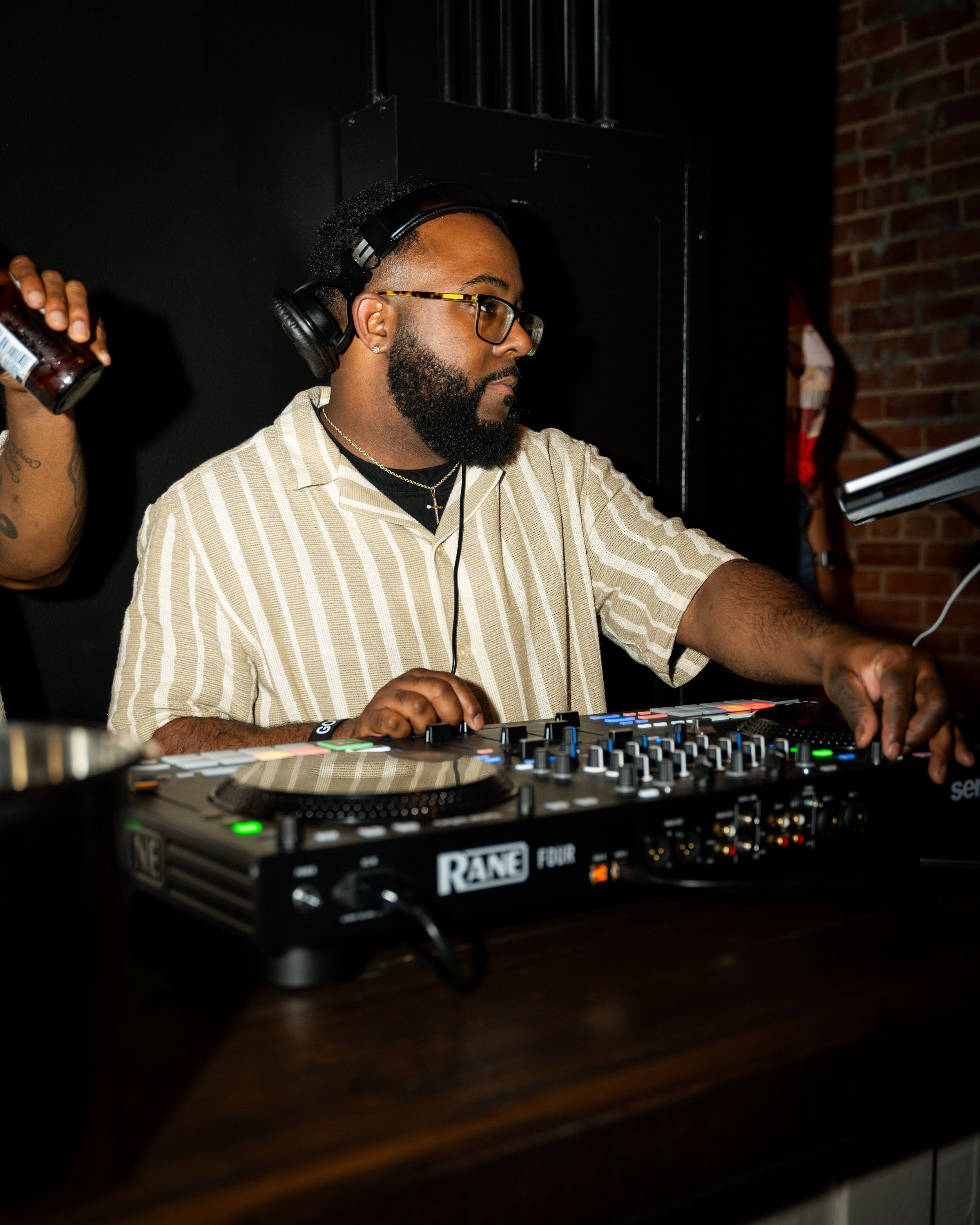 A DJ wearing glasses, headphones, and a striped shirt mixing music on a DJ controller with various controls and lights, in a dimly lit indoor venue with brick walls.
