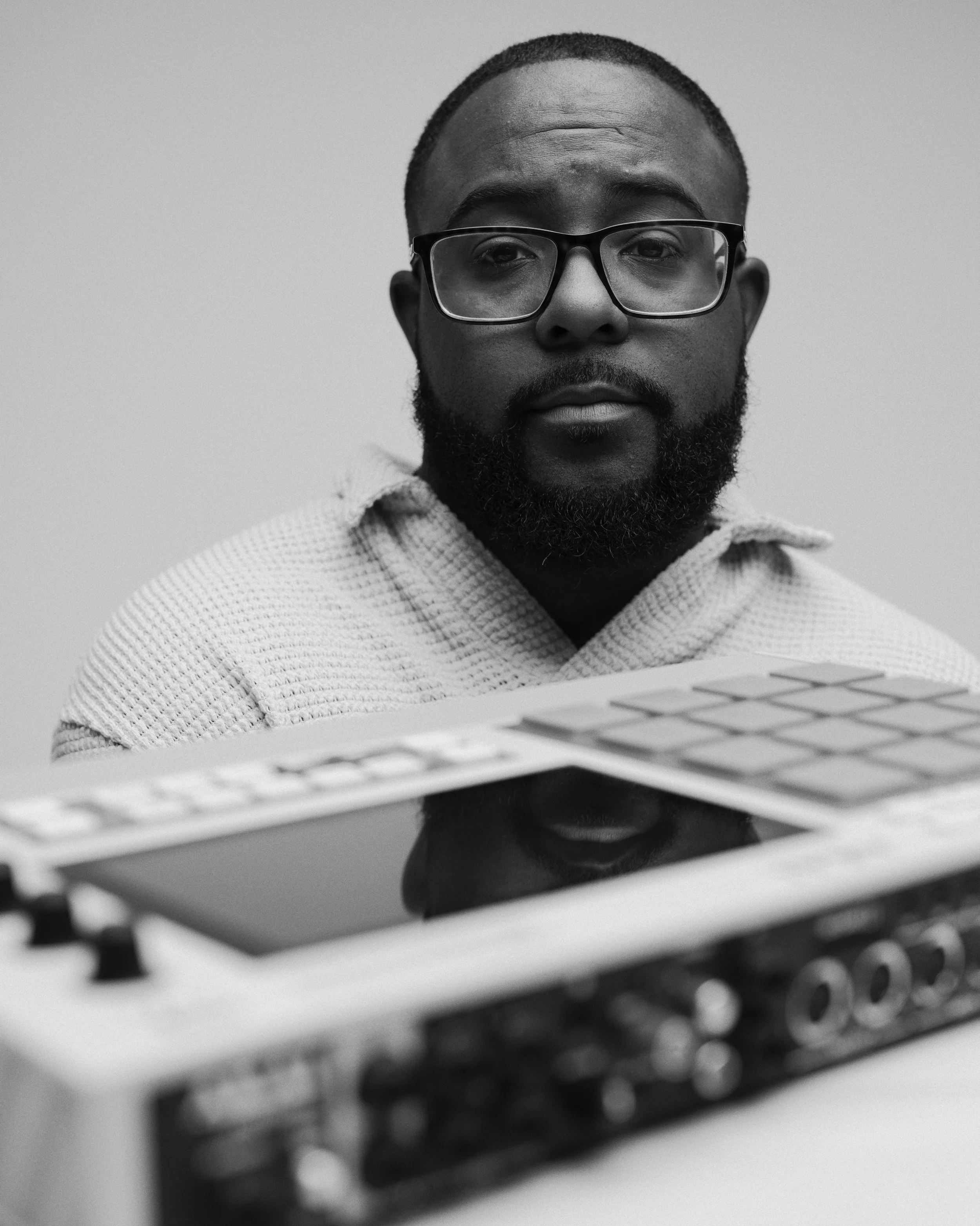 Black and white photo of a man with glasses, short hair, and a beard, looking at the camera, with a keyboard in the foreground.