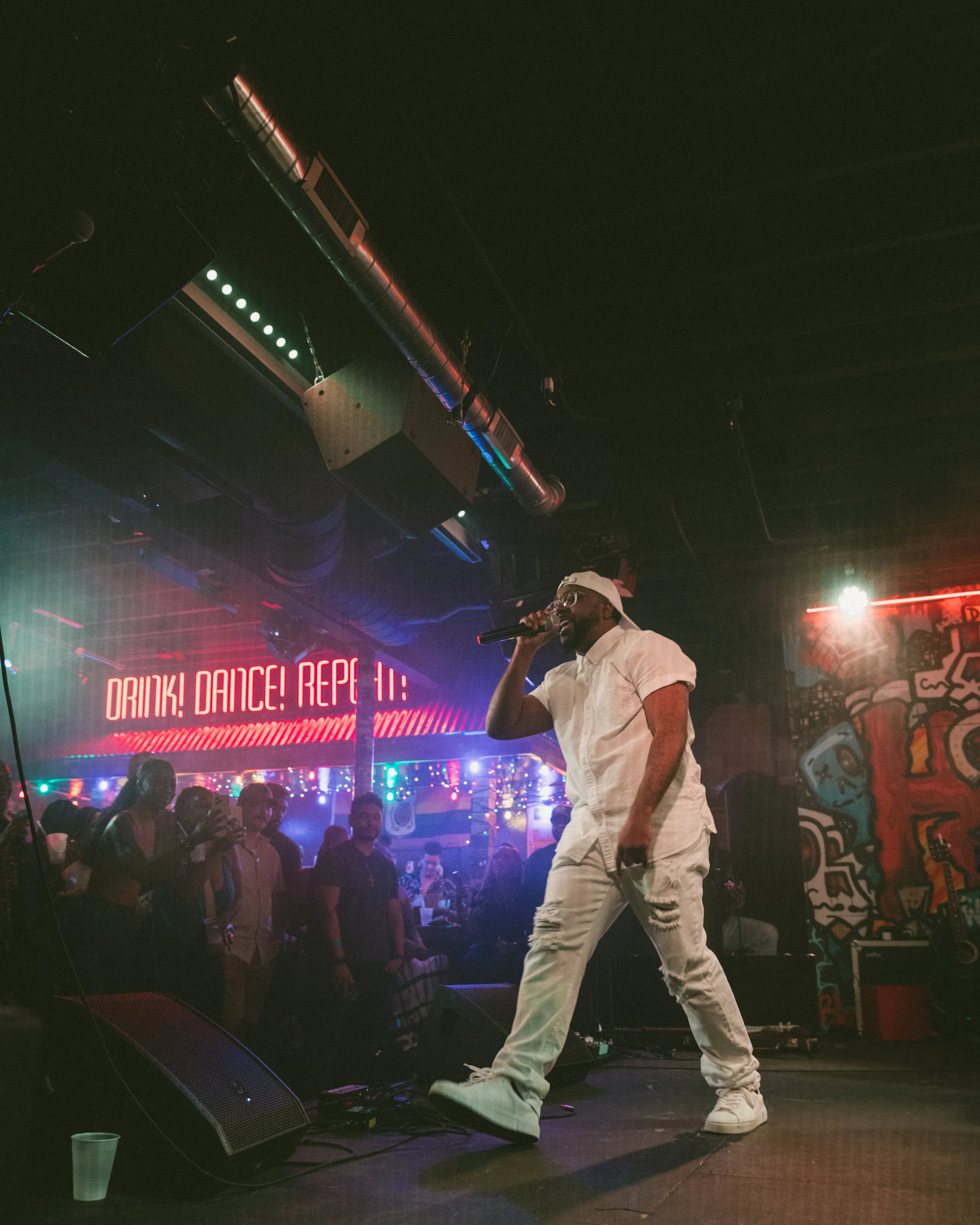 A man singing into a microphone on stage at a nightclub or bar, with a crowd of people watching him. Neon lights and wall decorations are visible in the background.
