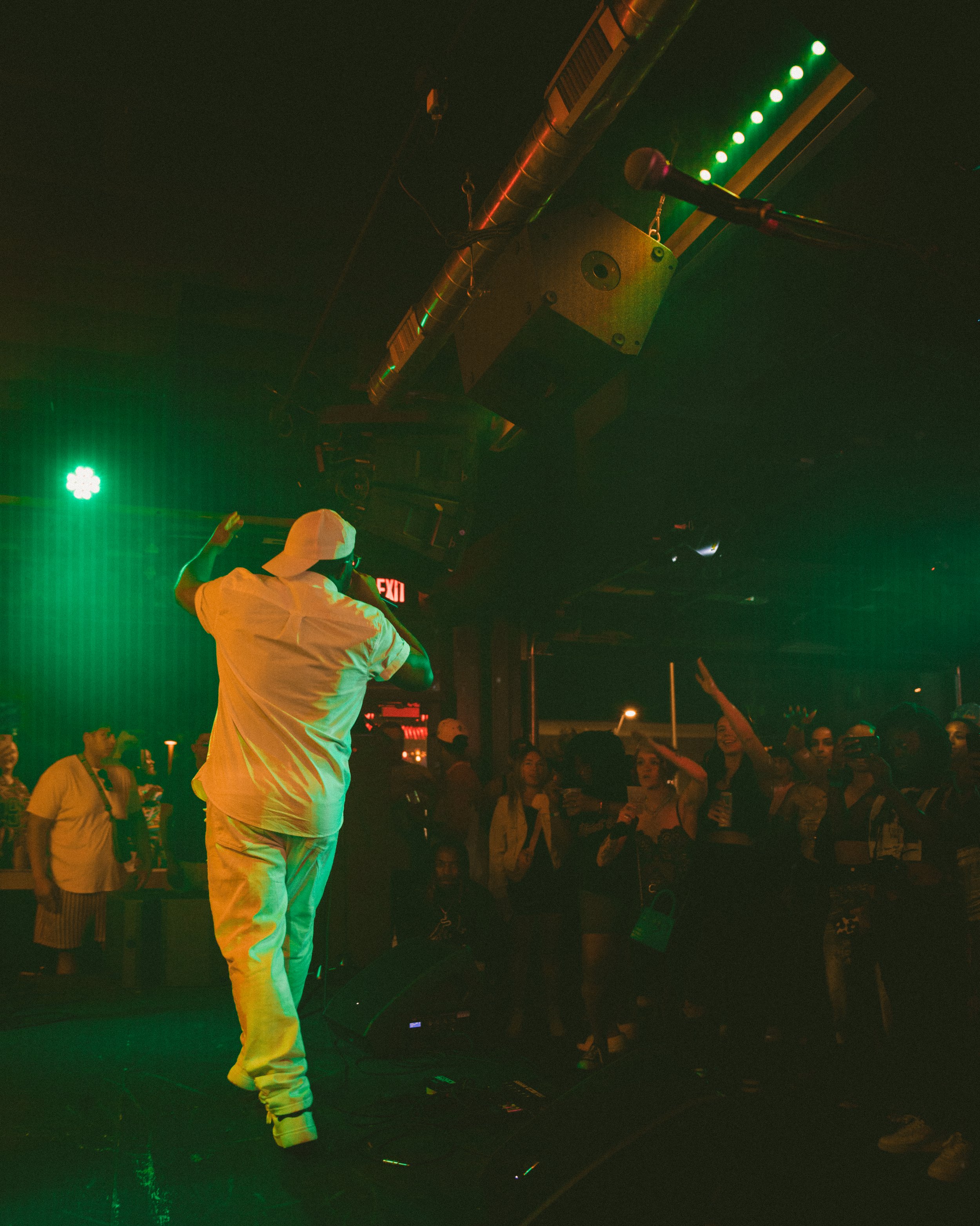 A person dressed in white with a hoodie, holding a microphone, performs on stage under green and warm stage lighting. Audience members are visible, some raising their hands, in a dark indoor setting with an exit sign