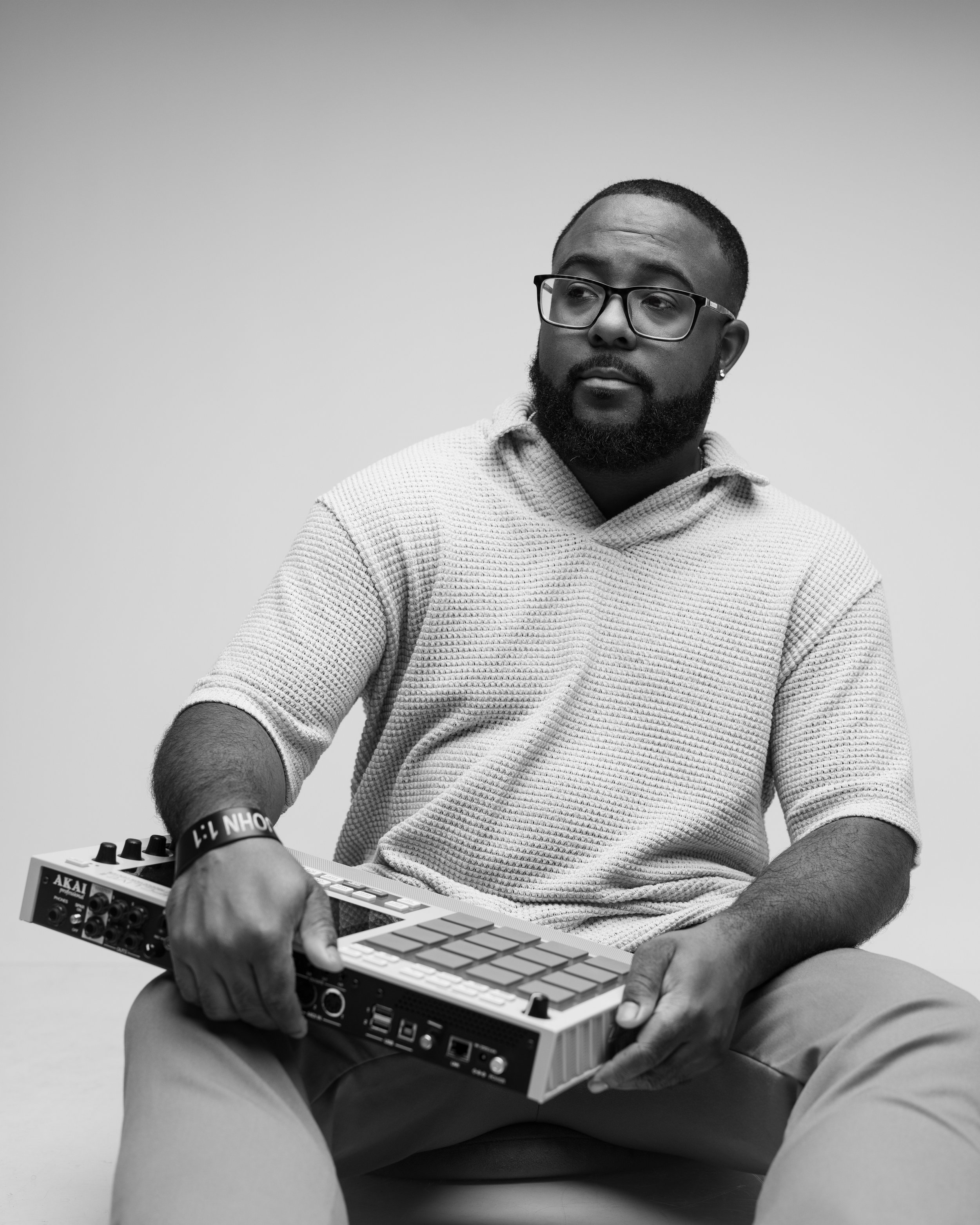 A young man with glasses and a beard is sitting on the floor, playing a MIDI controller keyboard. He is wearing a textured hoodie, shorts, and a wristband. The background is plain and light-colored.